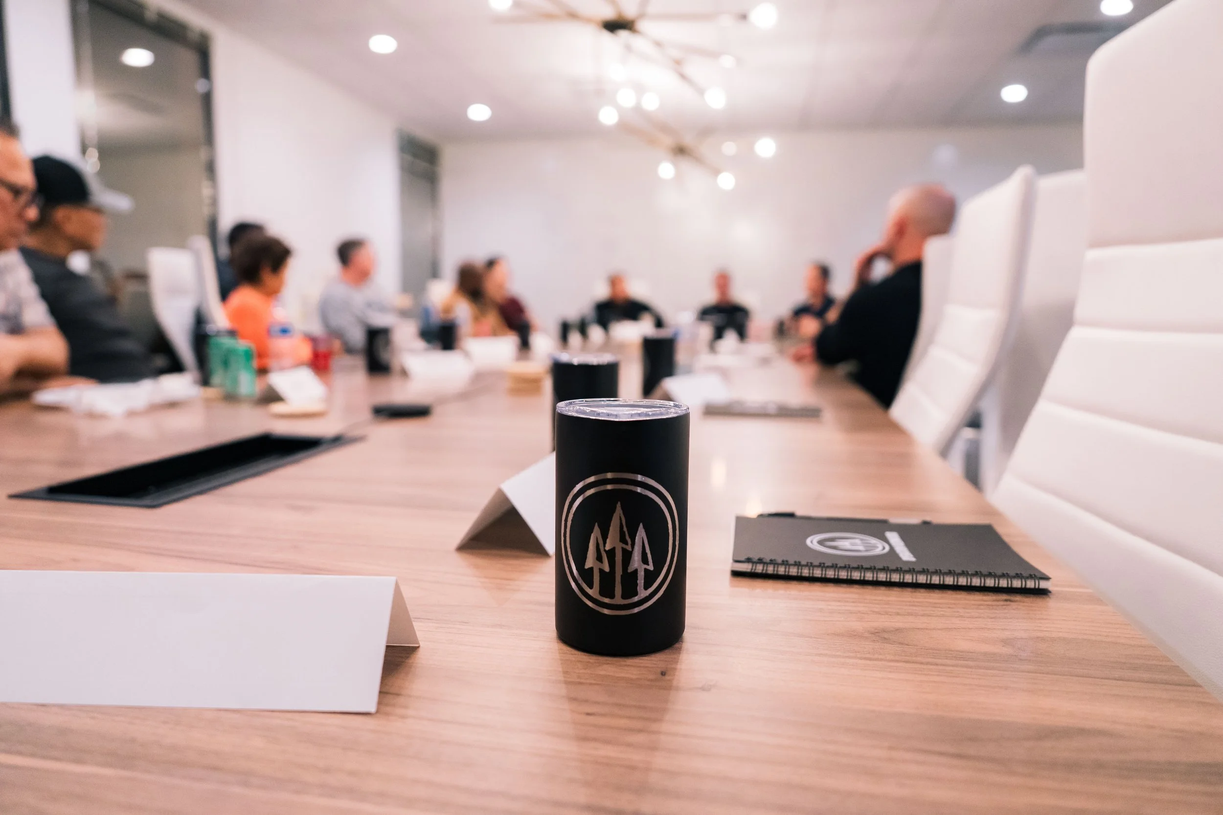 A meeting room with a long wooden table, white chairs, and a group of people seated around it. The table has a black cylindrical object with a logo, a spiral notebook, and several other items. The room is well-lit with ceiling lights.