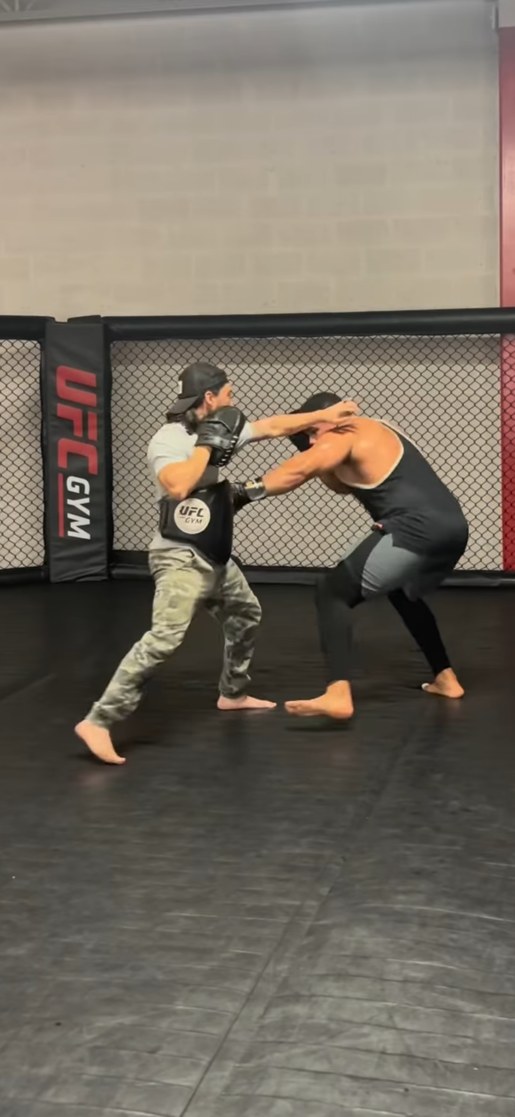 Dos hombres practicando pelea de entrenamiento en una jaula de MMA, uno con equipo de protección golpeando al otro.