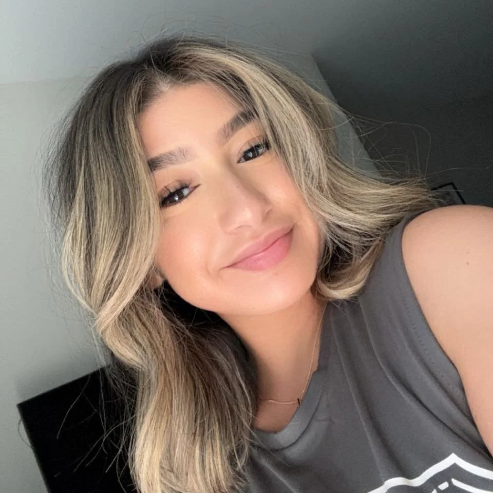 Close-up of a smiling woman with wavy blonde hair, wearing a gray top, indoors with a background wall and picture frame.
