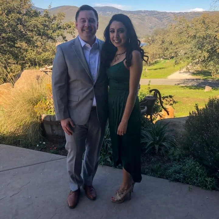 A man and woman standing outdoors, both dressed in formal attire, smiling with mountains and greenery in the background.