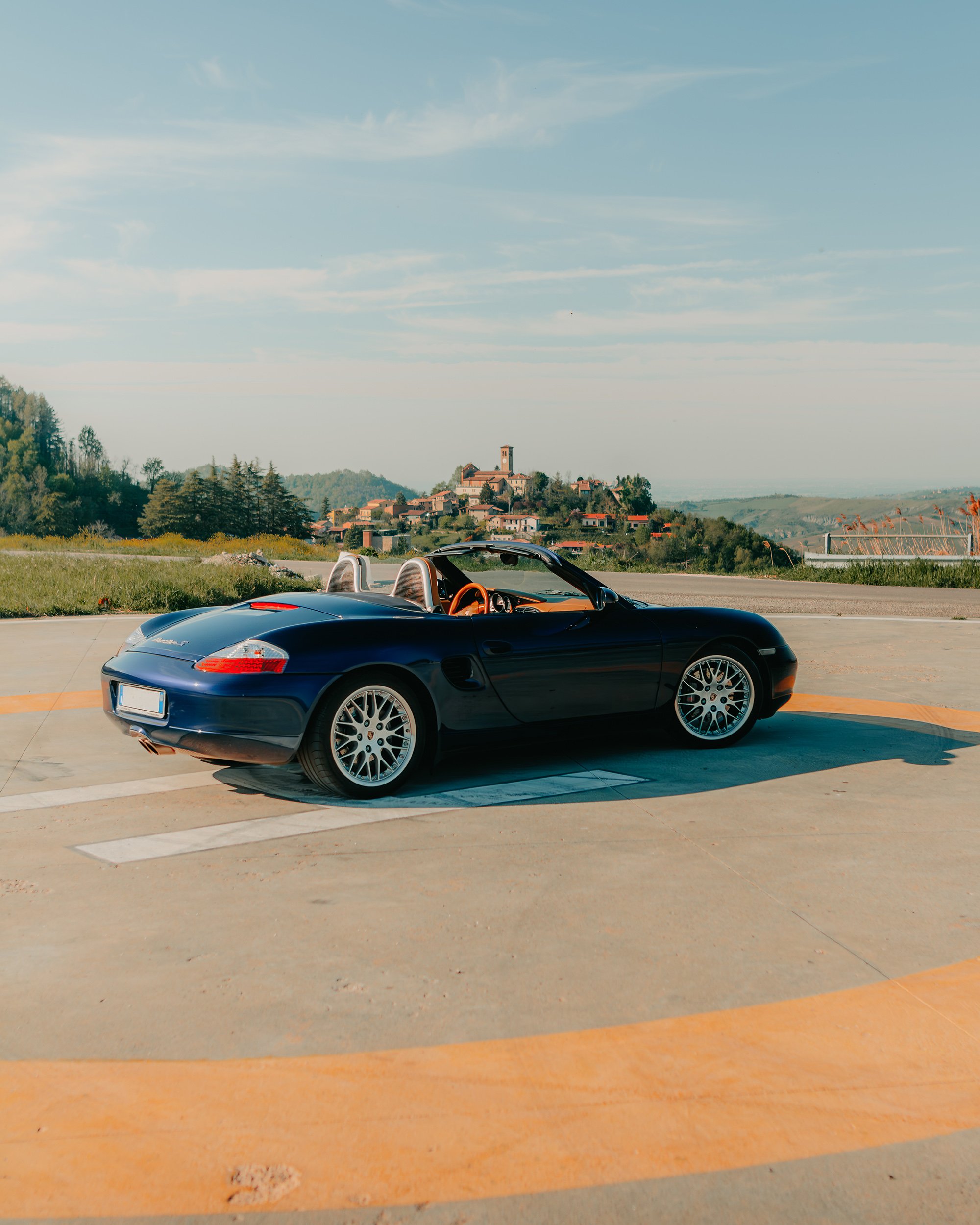 Auto sportiva blu decappottabile su strada panoramica, sullo sfondo un villaggio collinare.