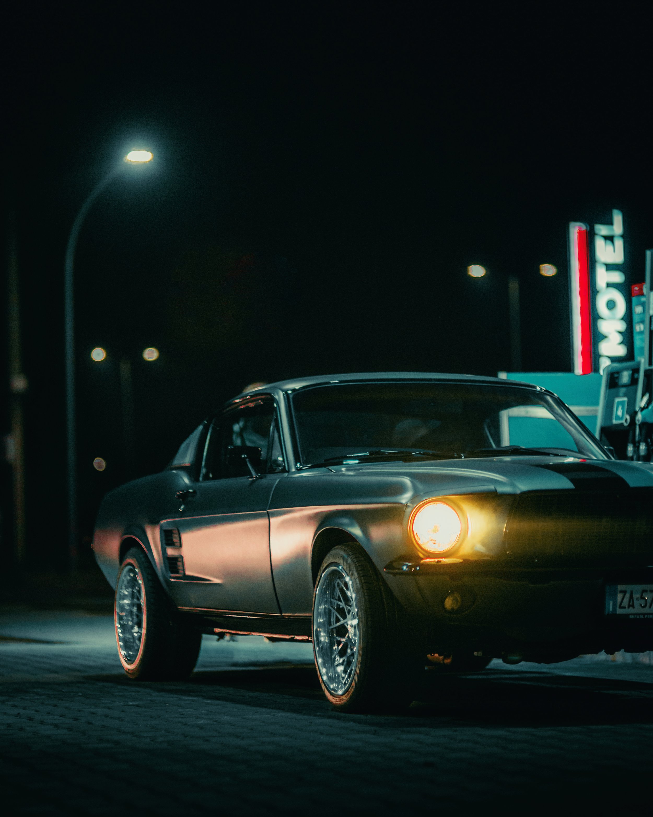 Auto classica nera presso una stazione di servizio di notte con insegna "MOTEL" sullo sfondo.