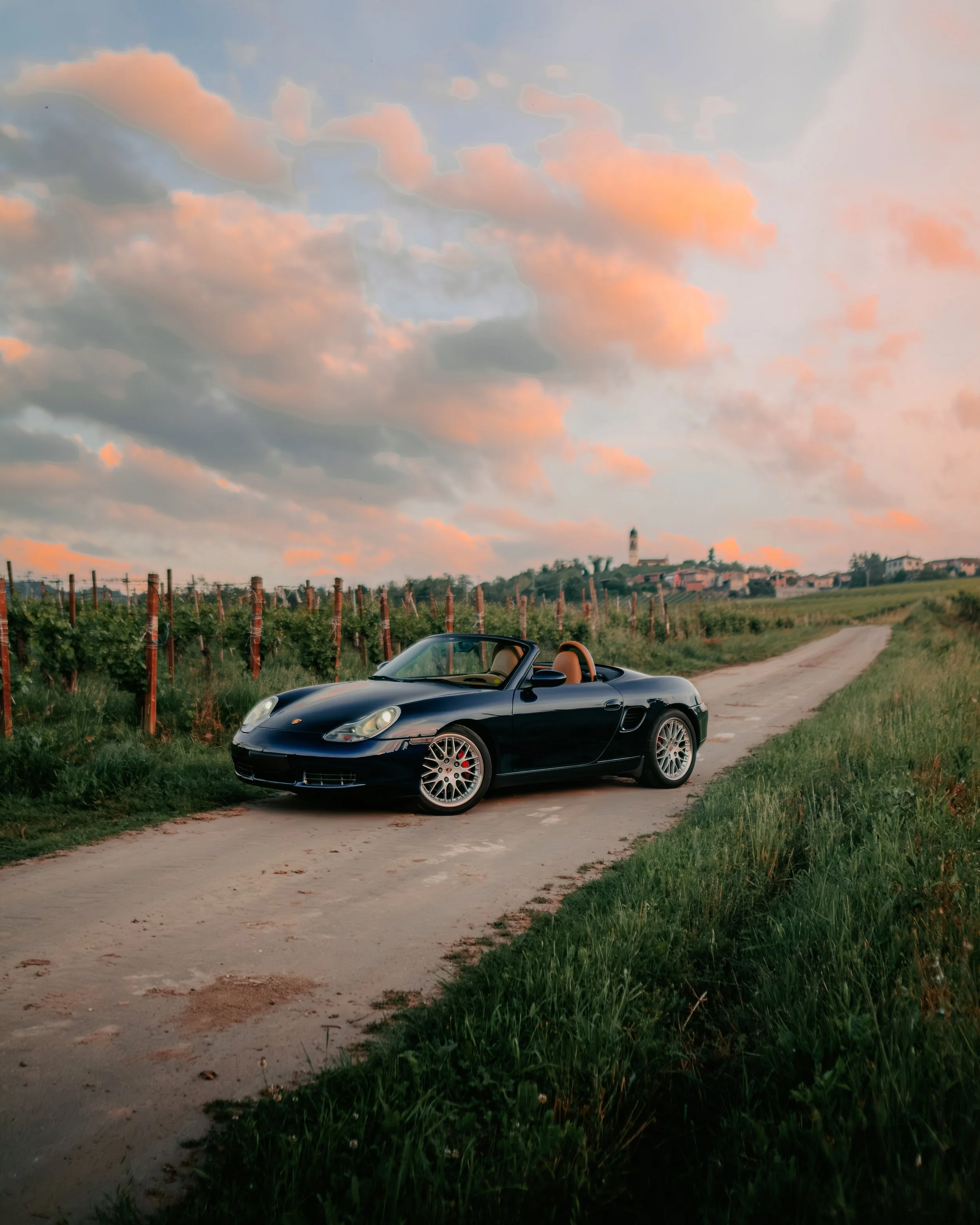 Auto sportiva decappottabile nera su strada di campagna con vigneti e cielo nuvoloso al tramonto.