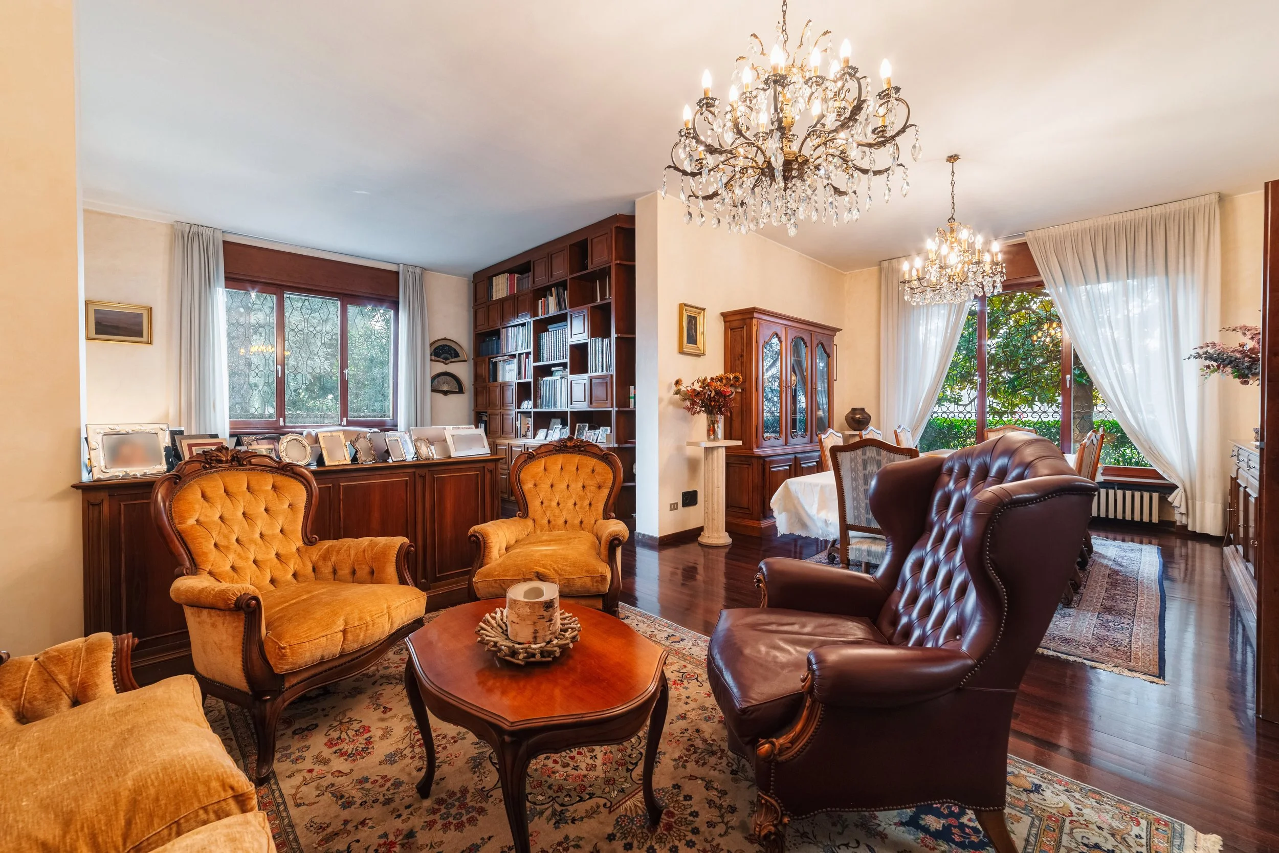 Soggiorno elegante con poltrone in pelle e velluto, tavolino in legno, libreria, lampadario di cristallo e finestre grandi con tende bianche, vista su giardino esterno.