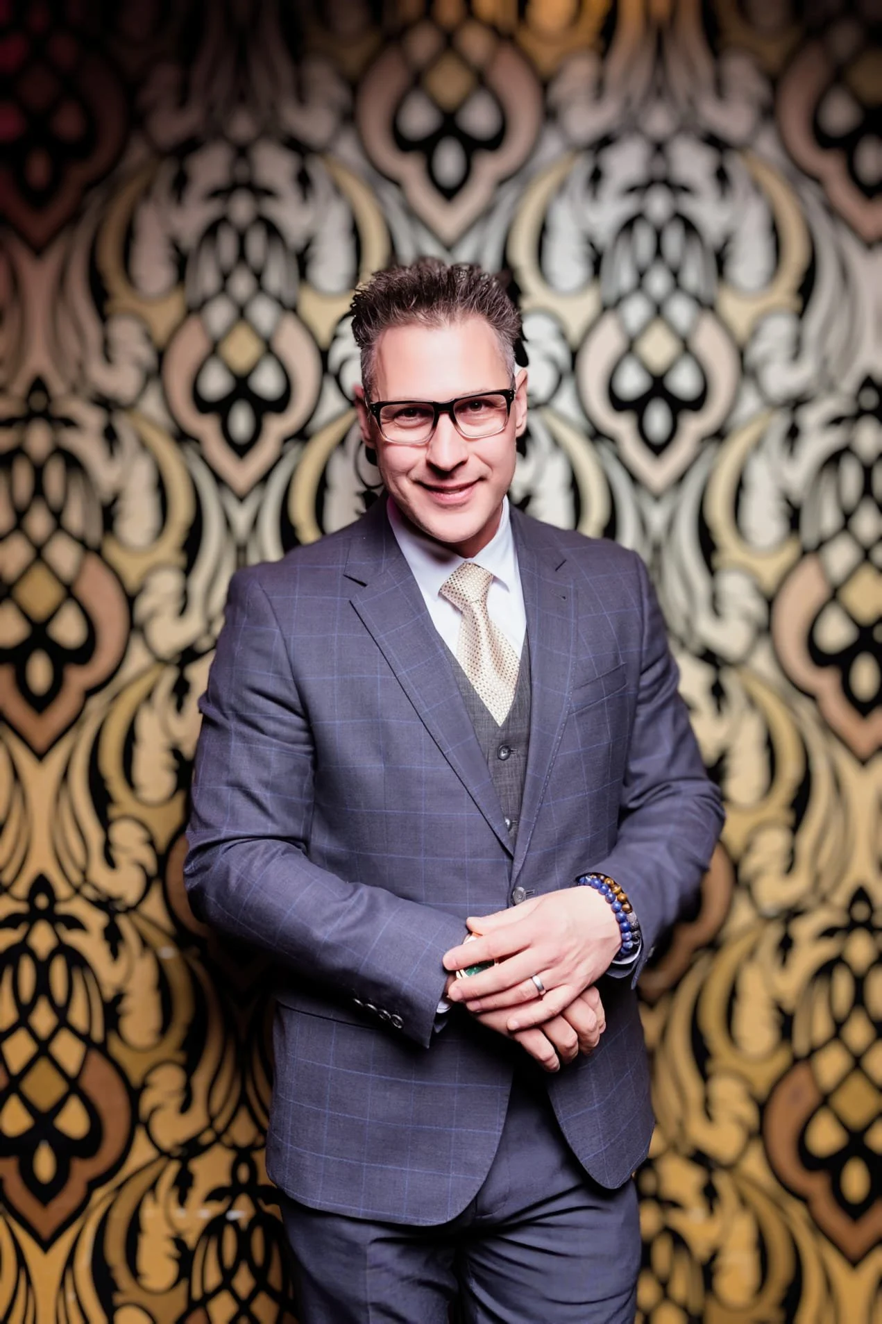 Man wearing glasses in a suit and tie, standing in front of a gold and black patterned wallpaper.