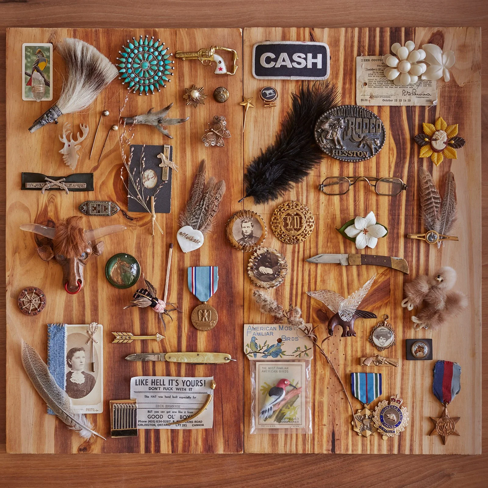 A wooden board displaying an assortment of eclectic vintage items including patches, pins, medals, feathers, postcards, small figurines, old photographs, a knife, eyeglasses, and various trinkets. Some notable items are a bull's head, a black feather, wooden carvings, and a card labeled 'CASH.'"