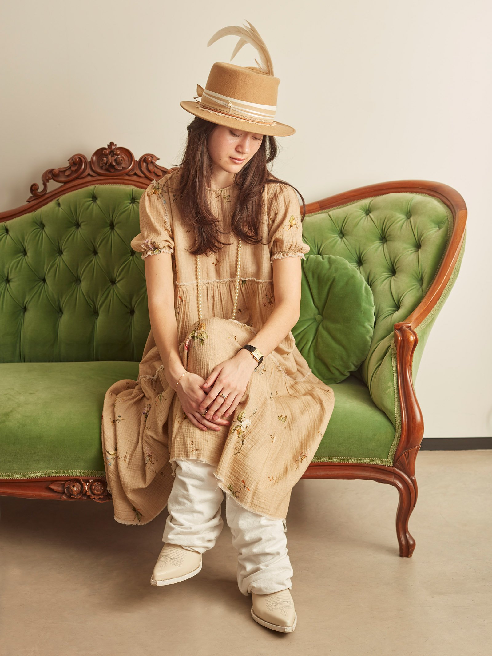 Person wearing a beige dress with floral patterns, a tan hat with feathers, and white boots, sitting on a vintage green velvet couch.