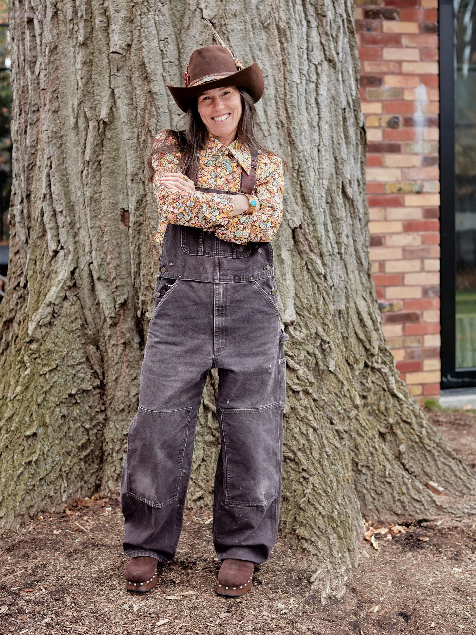 Person wearing a cowboy hat, floral shirt, and overalls standing in front of a tree trunk and brick wall.