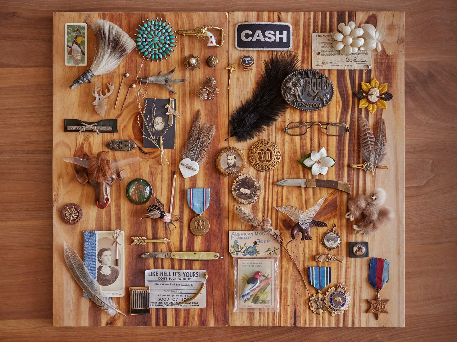 A wooden board displaying a collection of nostalgic and vintage items, including a feather, various pins, a cowboy hat, a pocket knife, old photographs, a cash patch, medals, and decorative objects like a bull's head and floral pieces.