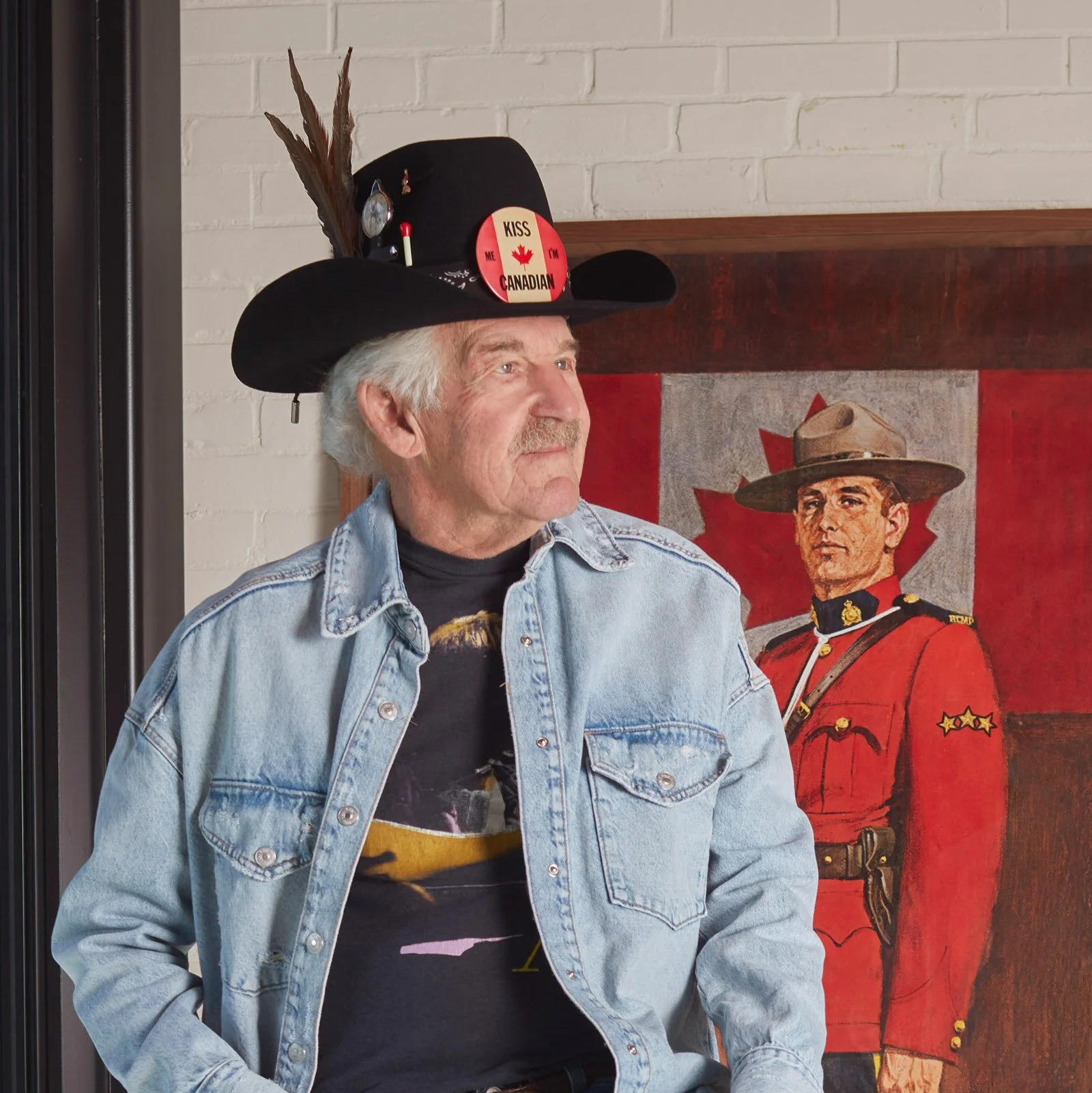 Man wearing a denim jacket and a hat with a "Kiss Me I'm Canadian" button, standing in front of a painting of a Mountie with a Canadian flag.