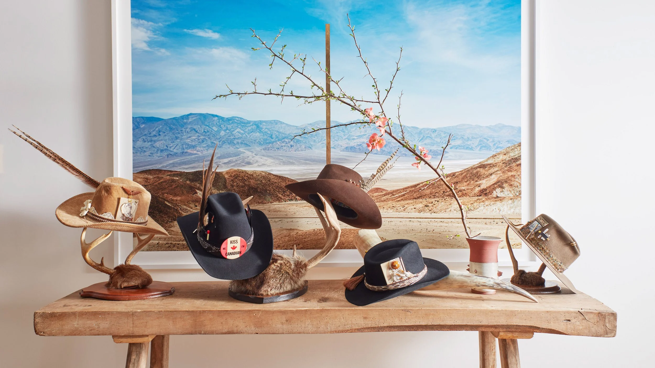 Decorative hats displayed on antler stands atop a wooden table with a scenic desert landscape photograph in the background.