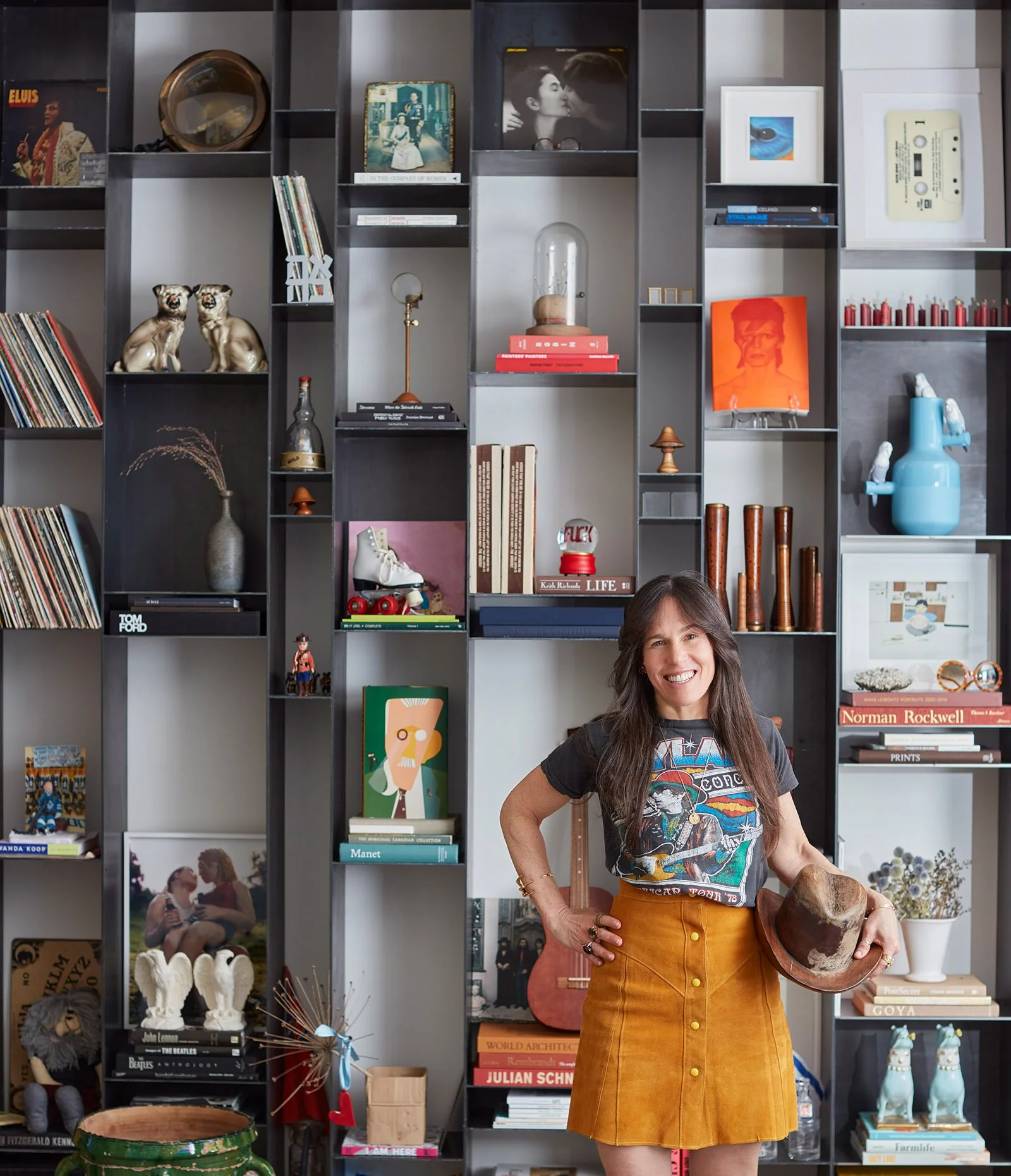 A woman with long hair stands in front of a large, eclectic bookshelf. The shelves are filled with a variety of items including vinyl records, books, figurines, artworks, and decorative objects like a blue pitcher and gold lion figurines. She is wearing a graphic t-shirt and a mustard yellow skirt, holding a brown hat in her hand, and smiling at the camera.