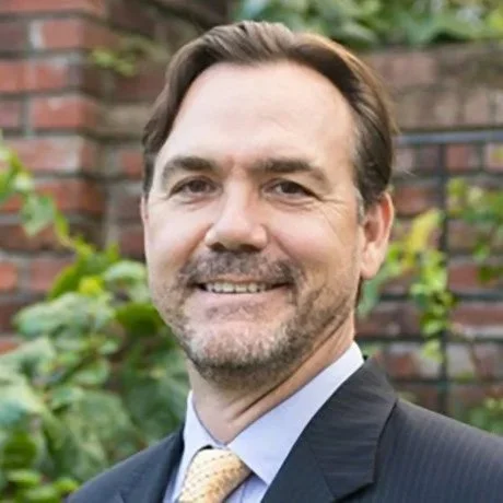 A middle-aged man with brown hair and a beard, smiling, wearing a dark suit, light blue shirt, and yellow tie, standing outdoors in front of a brick wall and green foliage.