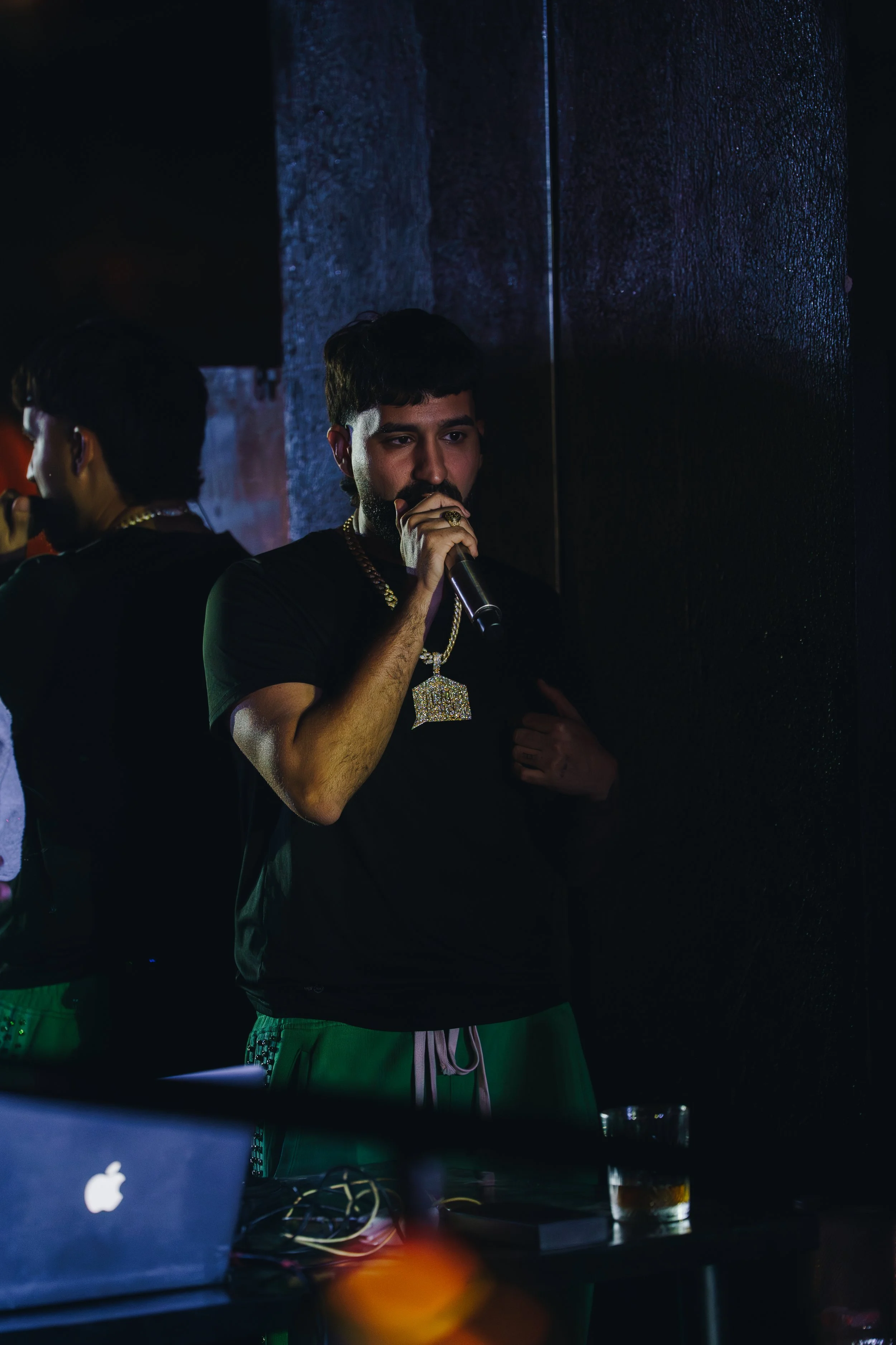 A man with a beard and short dark hair holding a microphone, standing next to a mirror, wearing a gold chain and a large diamond pendant necklace, in a dimly lit room; another person is partially visible in the mirror.