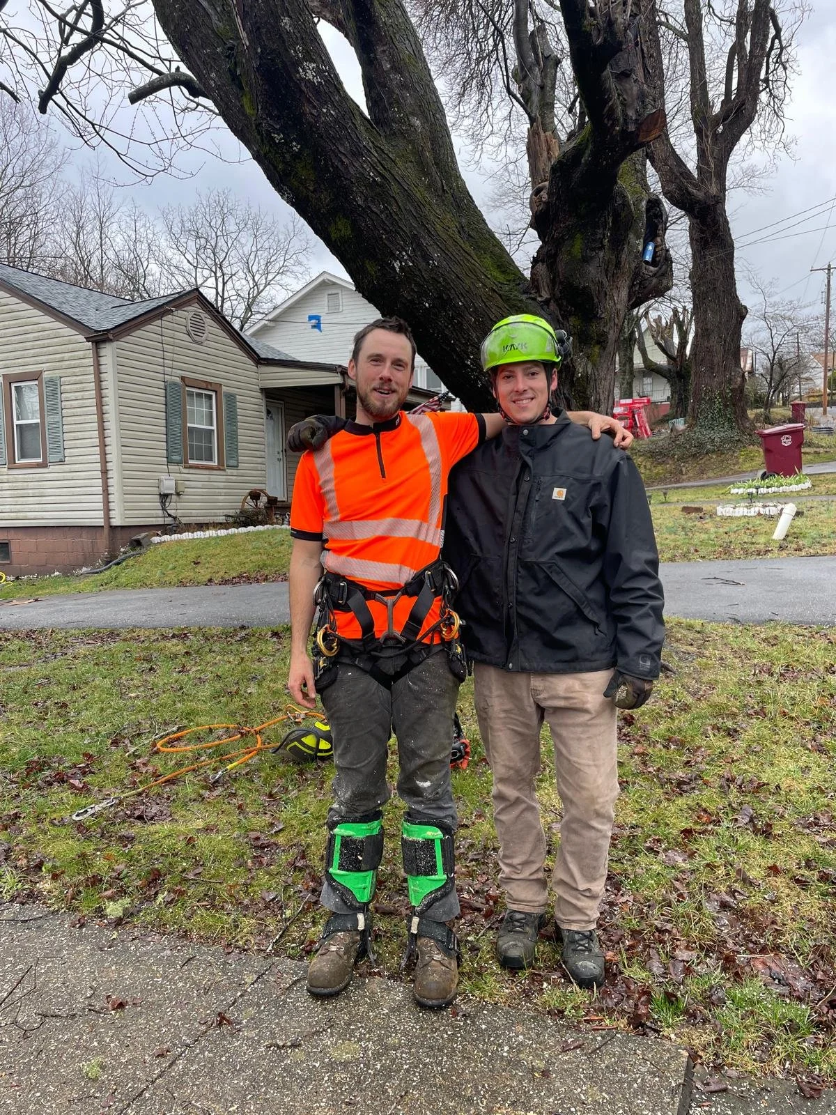 Two men standing outdoors in front of a large tree, smiling and posing with their arms around each other. One man is wearing a bright orange safety shirt, black gloves, and safety harness gear, while the other is in a black jacket and a green safety helmet. The background shows a residential neighborhood with houses, leafless trees, and a cloudy sky.