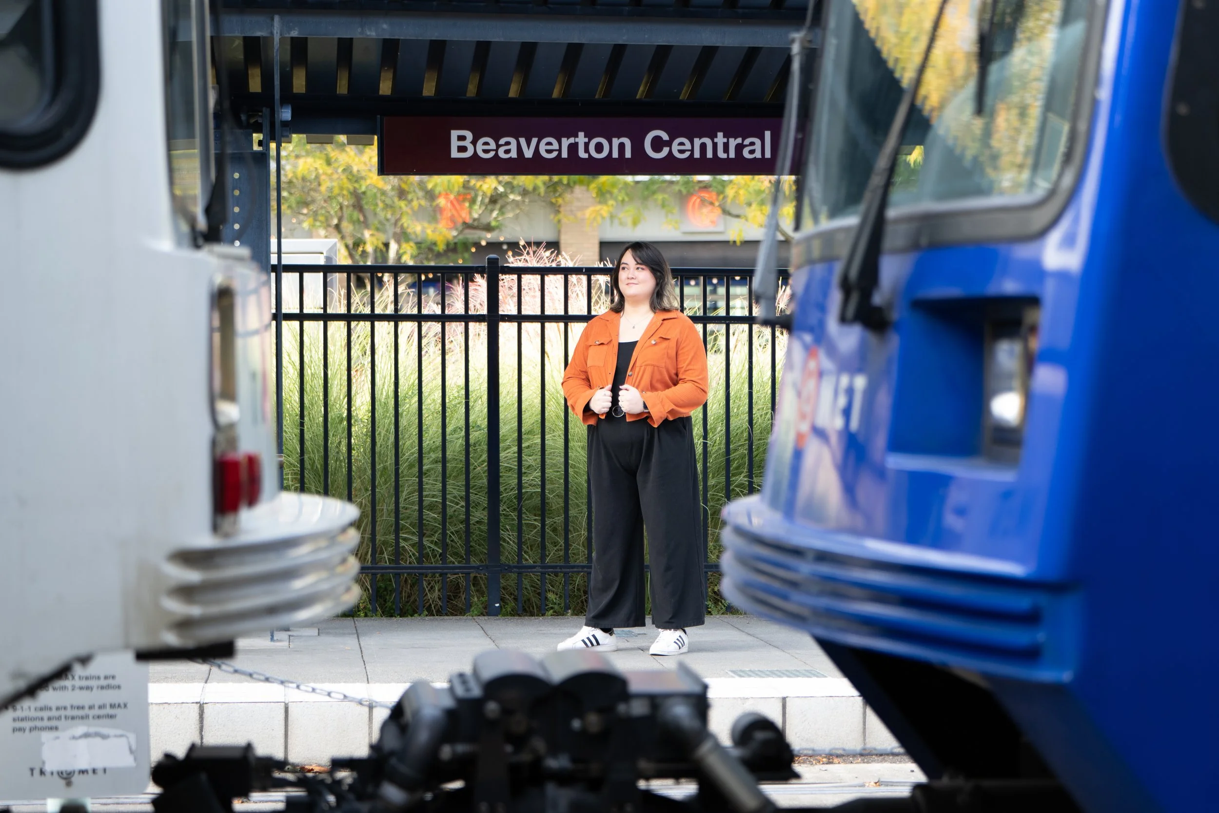 A woman in an orange jacket, black pants, and white sneakers standing at Beaverton Central transit station, looking to the side, between two MAX trains parked at the station.