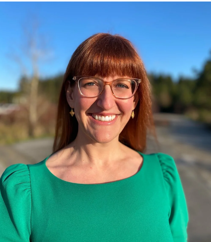A smiling woman with red hair, glasses, and wearing a green dress standing outdoors on a sunny day.