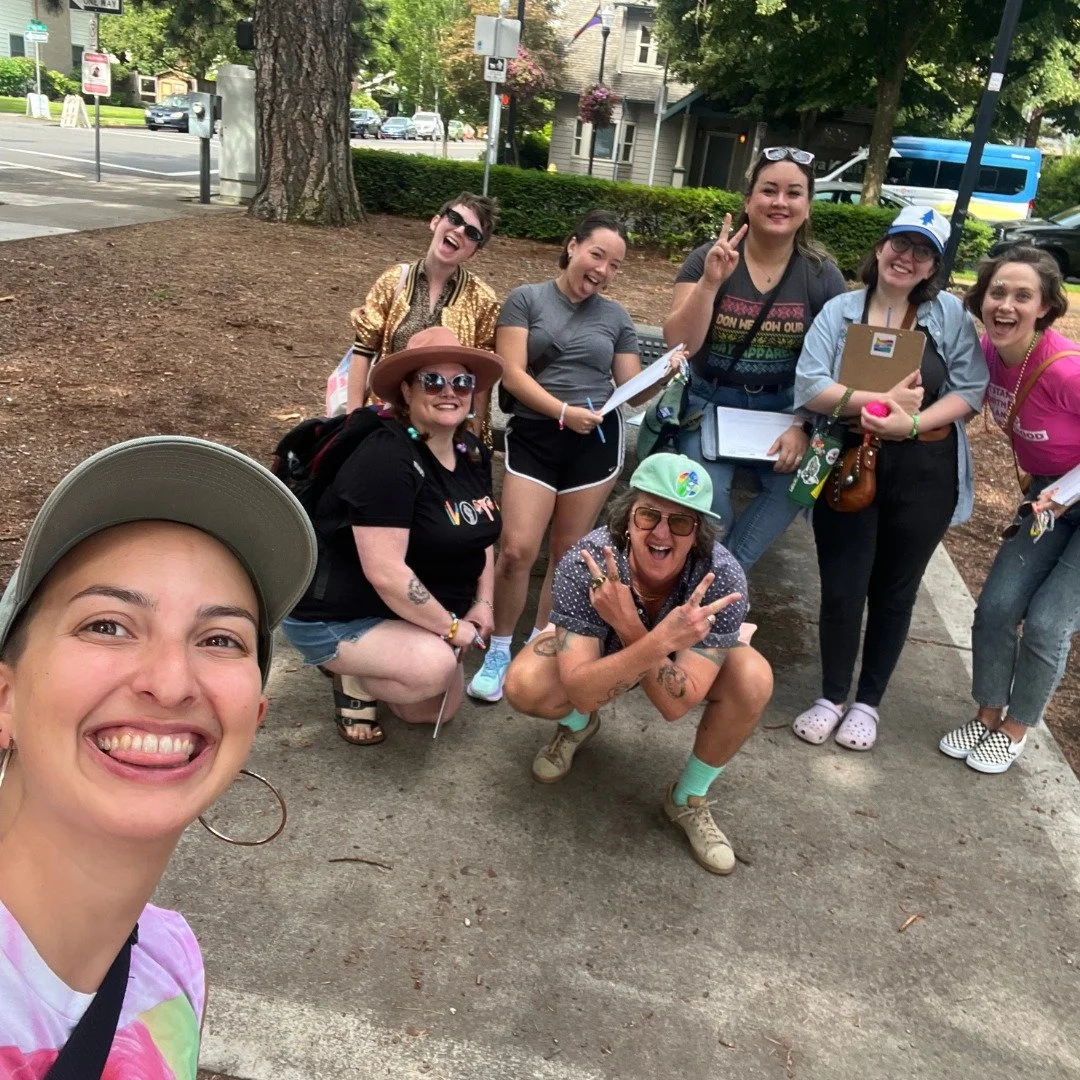 A diverse group of nine people posing for a photo on a picnic table in Beaverton City Park with trees in the background. They are smiling and making cheerful gestures, some holding clipboards.