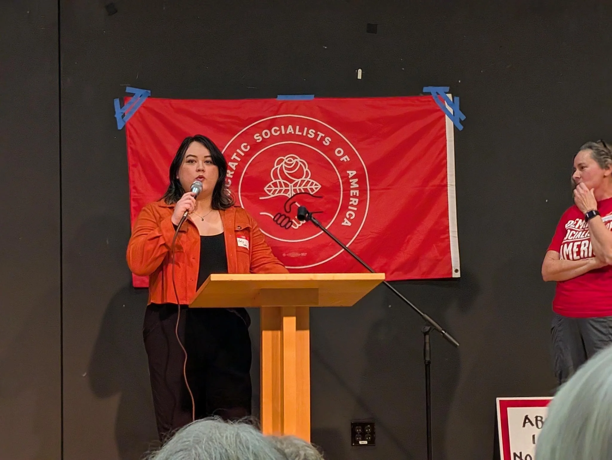 A woman in an orange jacket speaking at a podium with a microphone at a Democratic Socialists of America event, framed by a red banner with the DSA logo, standing next to another woman wearing a red shirt, on stage against a black background