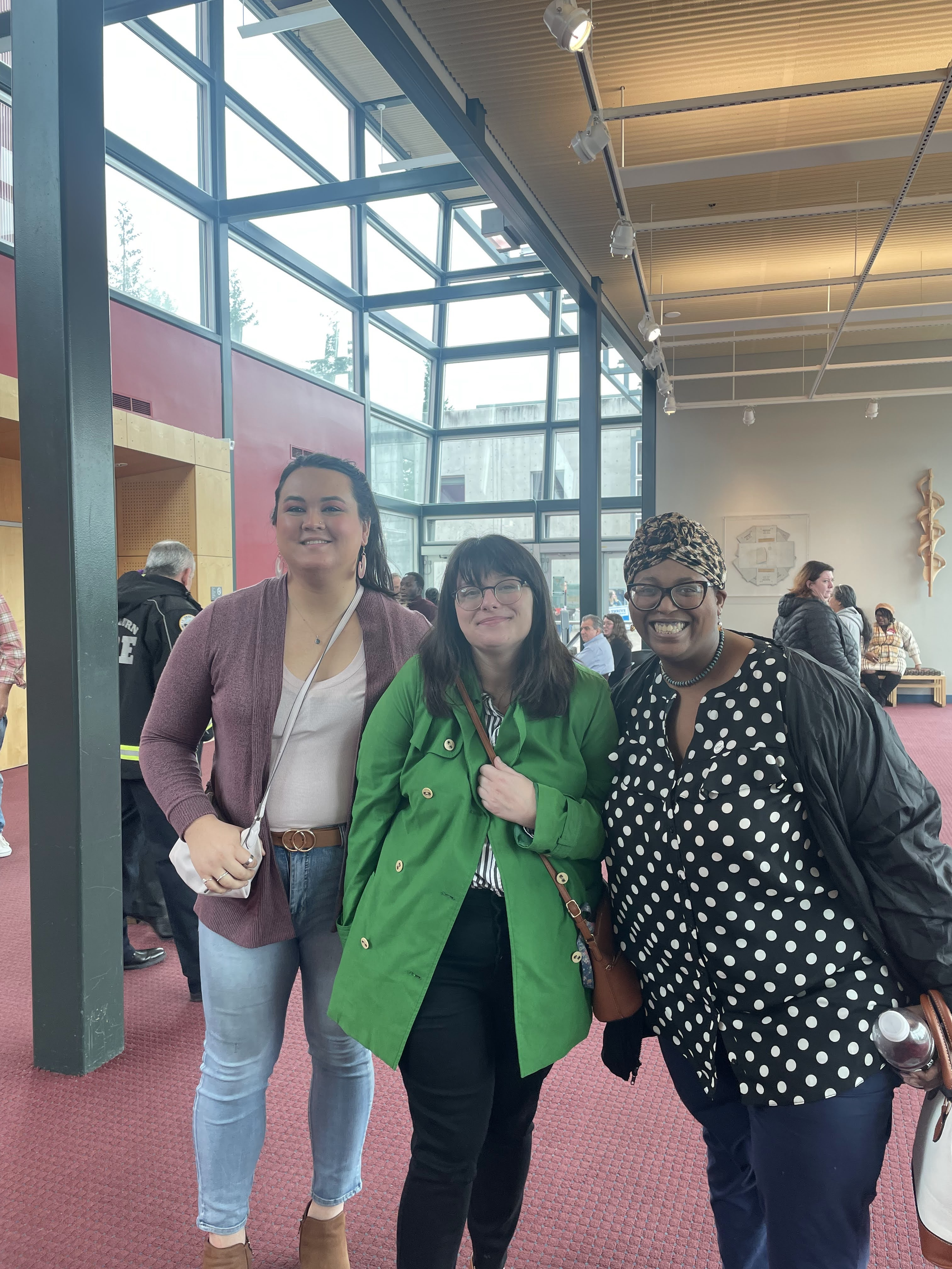 Three smiling women standing together indoors with large glass windows and a high ceiling, other people in the background.