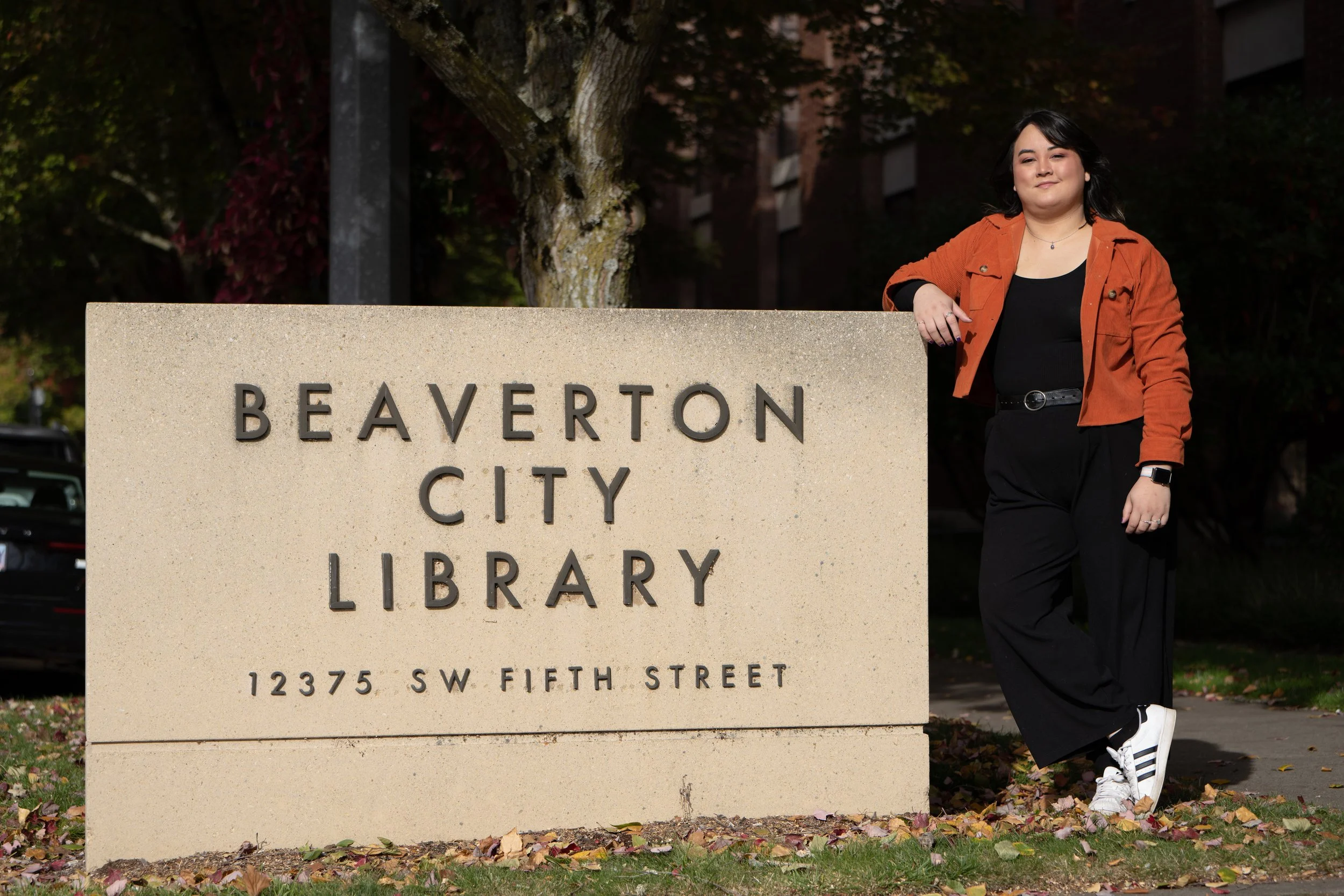 A woman standing next to a sign that reads 'Beaverton City Library, 12375 SW Fifth Street,' outdoors in autumn, wearing black pants, a black top, an orange jacket, and white sneakers.