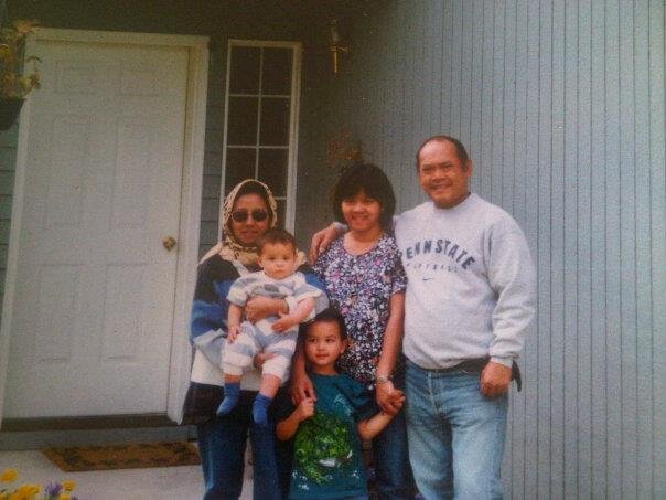 A family of five standing outside a house with an older woman wearing sunglasses and a hijab holding a baby, a young child, a young woman, and an older man.