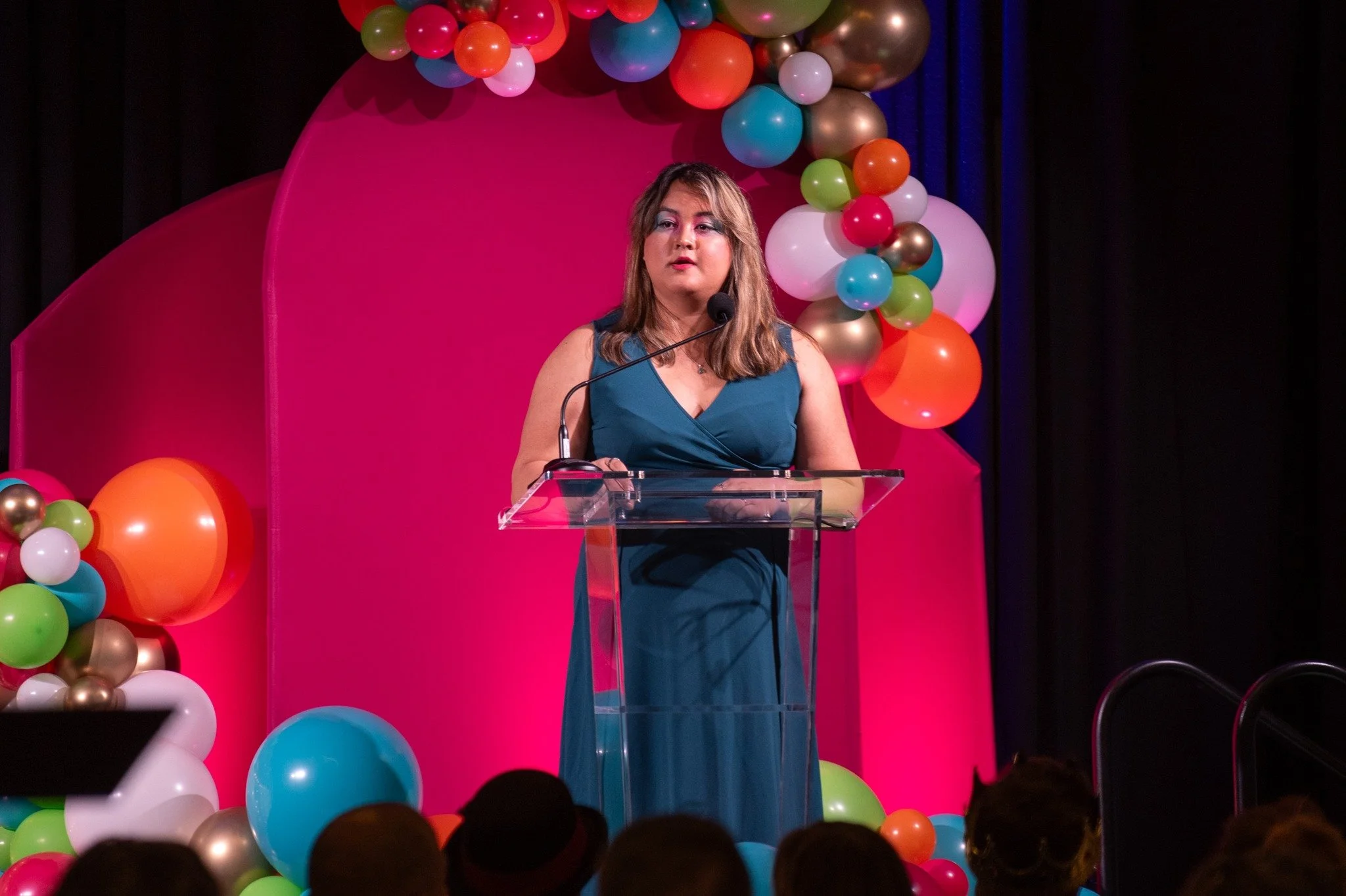 A woman in a sleeveless teal dress standing behind a clear podium giving a speech on stage decorated with pink background and colorful balloons.