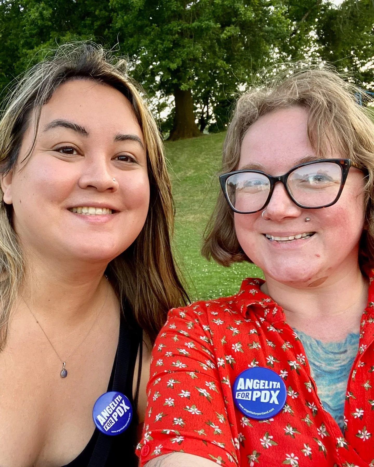 Two women outdoors smiling, wearing 'Angelita for Portland' campaign buttons, with trees and grass in the background.