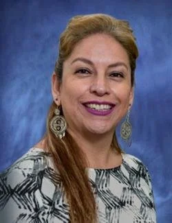 Portrait of a woman with light brown hair, earrings, and a patterned top against a blue background.