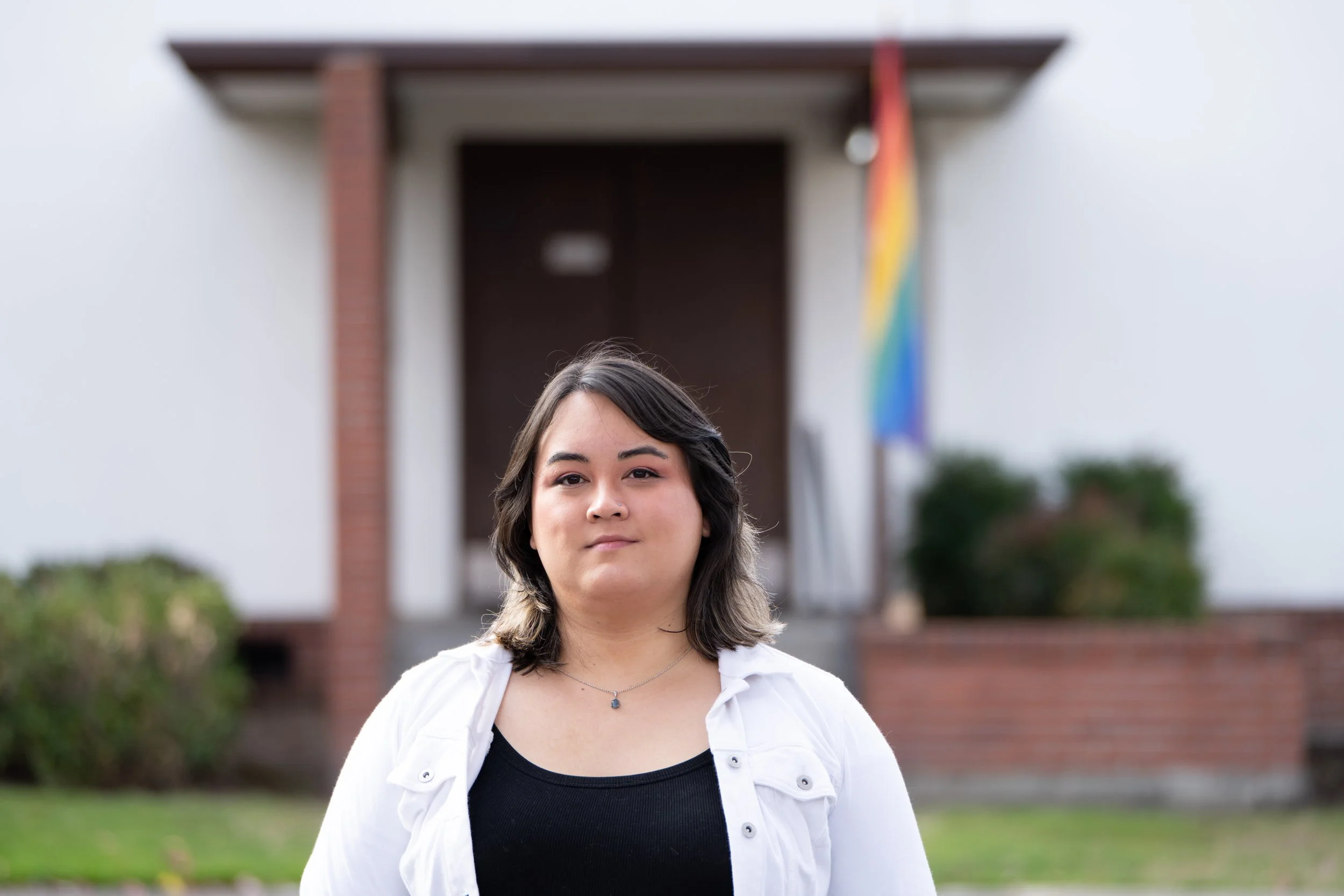 A young woman with shoulder-length dark hair, wearing a black top and white jacket, standing outdoors in front of a building with a rainbow flag on the right side.