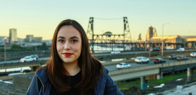 A young woman with dark hair and red lipstick standing outdoors with a bridge and cityscape in the background.