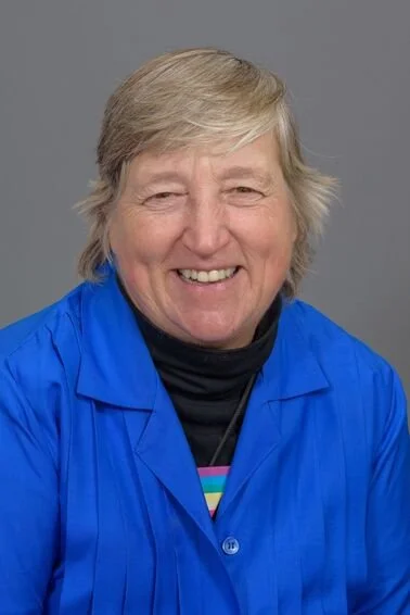 A smiling woman with short blond hair wearing a blue jacket and a black turtleneck, against a gray background.