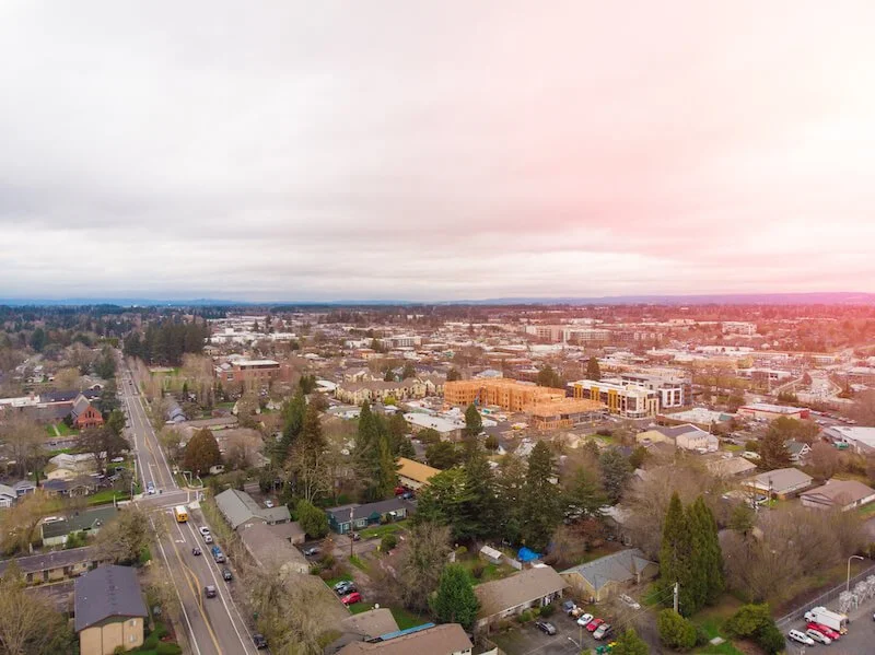 Aerial view of a suburban cityscape with residential houses, trees, roads, and some commercial buildings, under a cloudy sky with a pinkish hue from the sunset.