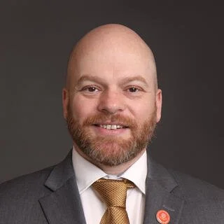 A bald man with a beard in a suit and tie, smiling against a dark background.