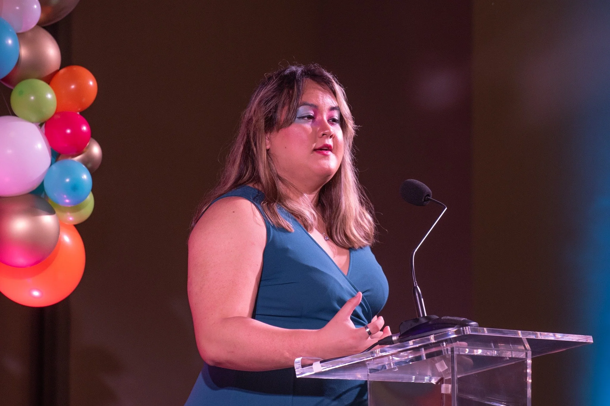 A woman with black hair standing at a clear acrylic podium with a microphone, wearing a sleeveless dark blue dress, in front of a background of colorful balloons.