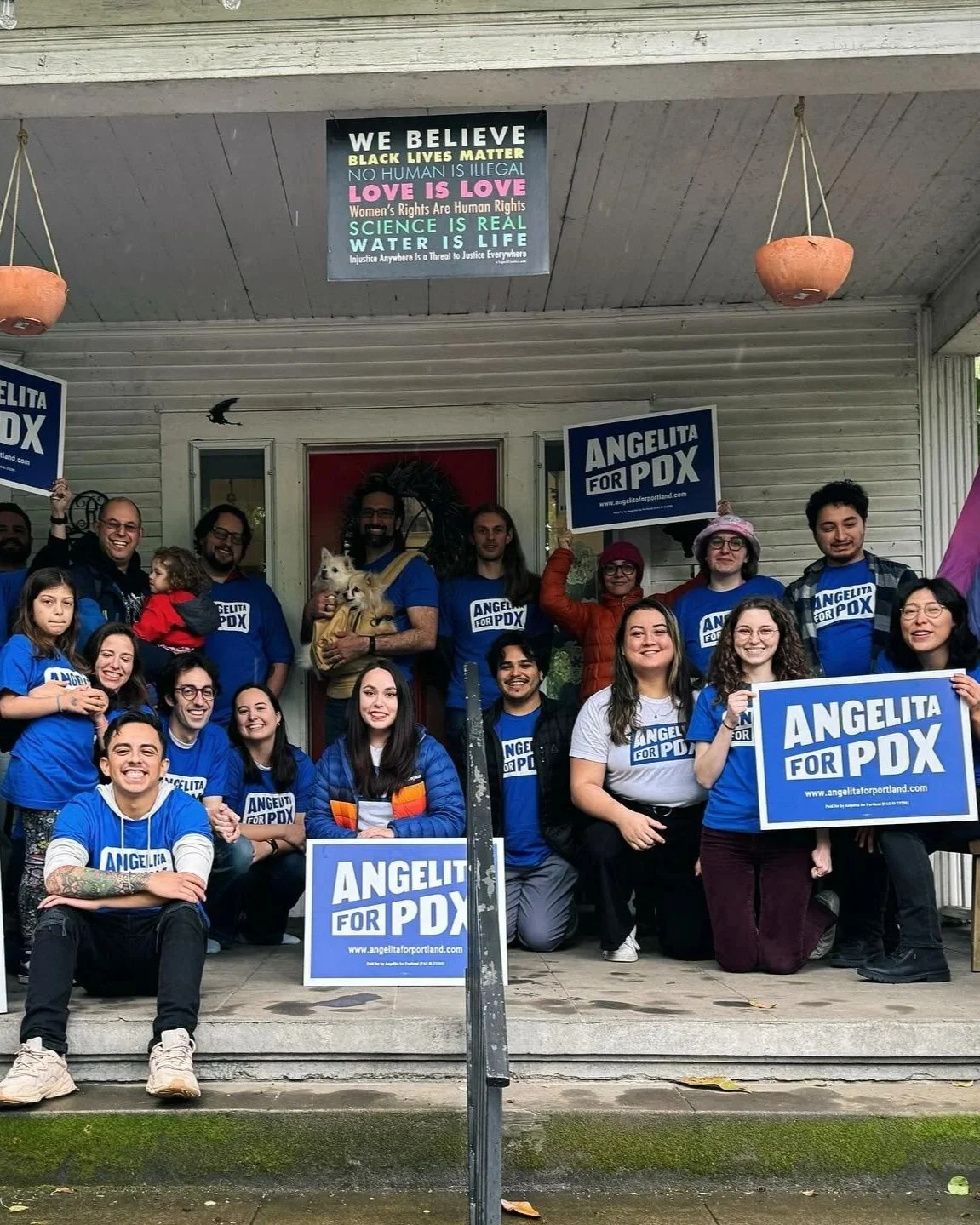 Group of people standing and sitting in front of a house, holding signs that read 'Angelita for PDX' and wearing blue shirts supporting Angelita's campaign. Some are smiling and posing for the photo, with two small dogs held by the person in the middle. A poster above them and a sign on the house also support Angelita for PDX.