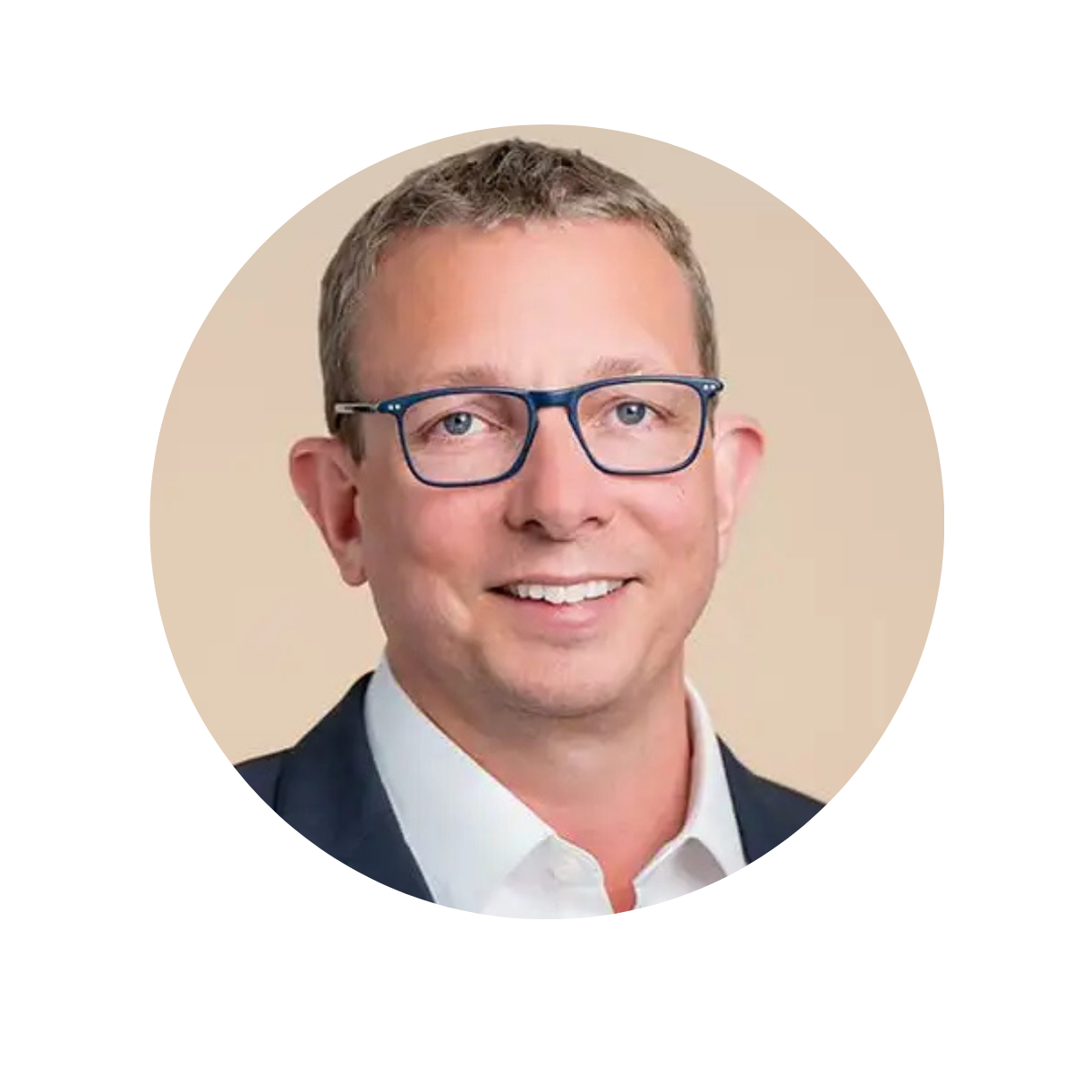 Headshot of a man with short light brown hair, wearing blue glasses, a white collared shirt, and a dark blazer, smiling against a beige background.