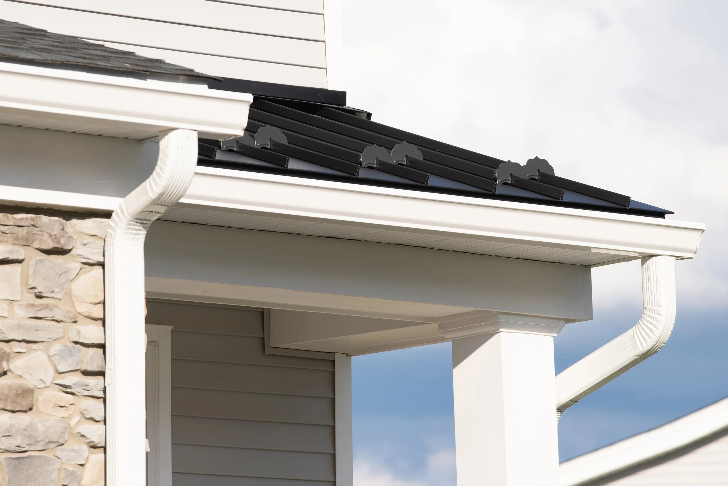 Close-up of white gutter system and black metal roof on a house with stone and vinyl siding exterior.