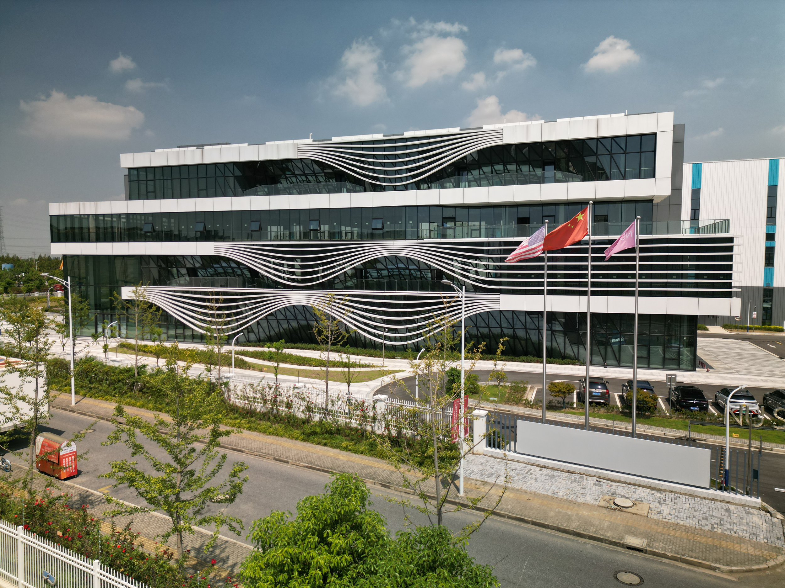 International OTC and personal care contract manufacturing facility supporting U.S.–China. Modern office building with wave-like white architectural features on the facade, four flags including the United States and China, and a parking lot in front.