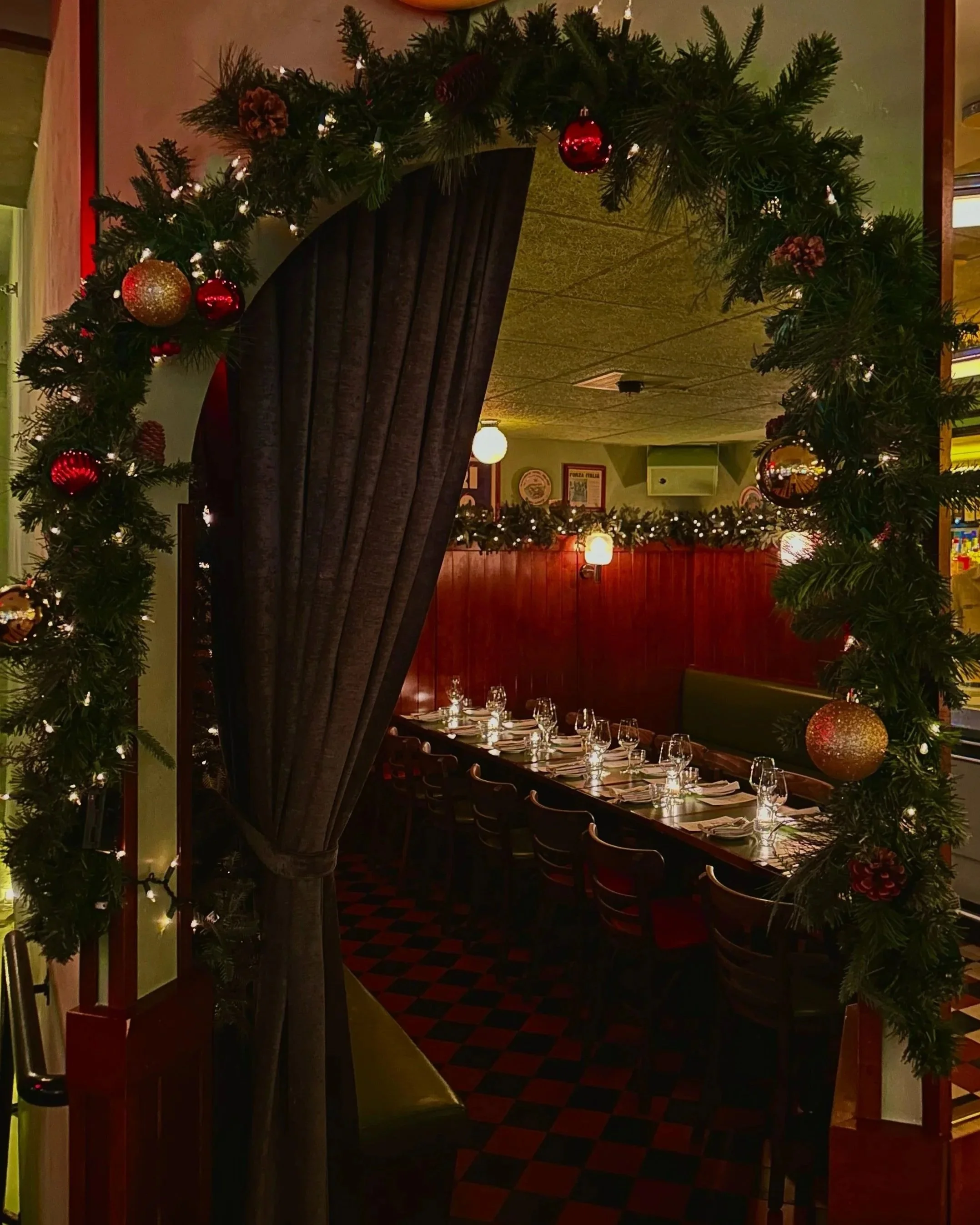 Festively decorated restaurant with a large table set for dining, adorned with glasses, plates, silverware, and napkins, framed by a green garland with red and gold ornaments and pinecones, with warm lighting and dark curtains.