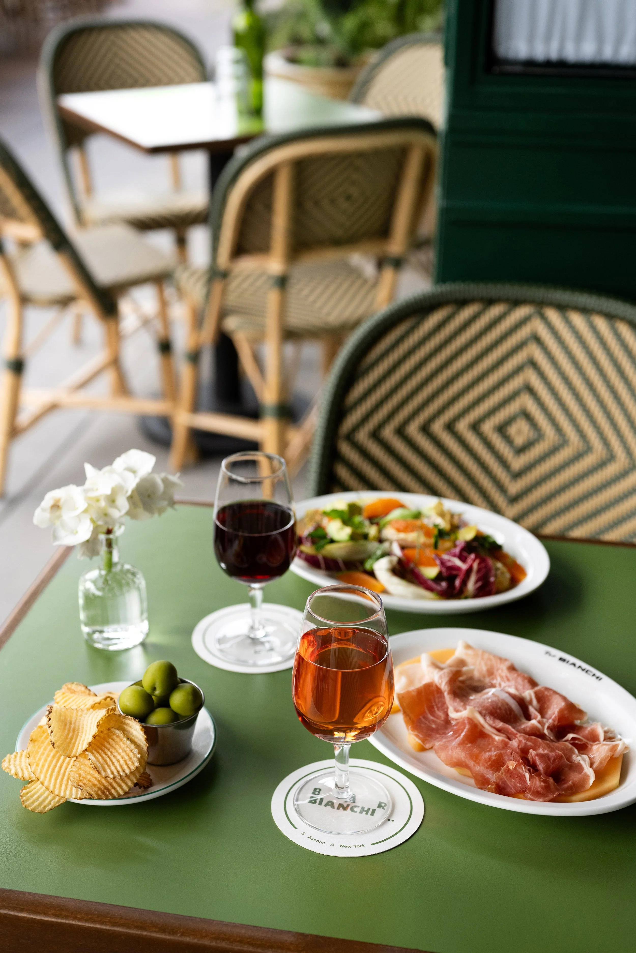 An outdoor dining table set with two glasses of red and rosé wine, a plate of prosciutto, a bowl of green olives, a side salad, and a small vase with white flowers, on a green table with rattan chairs in the background.