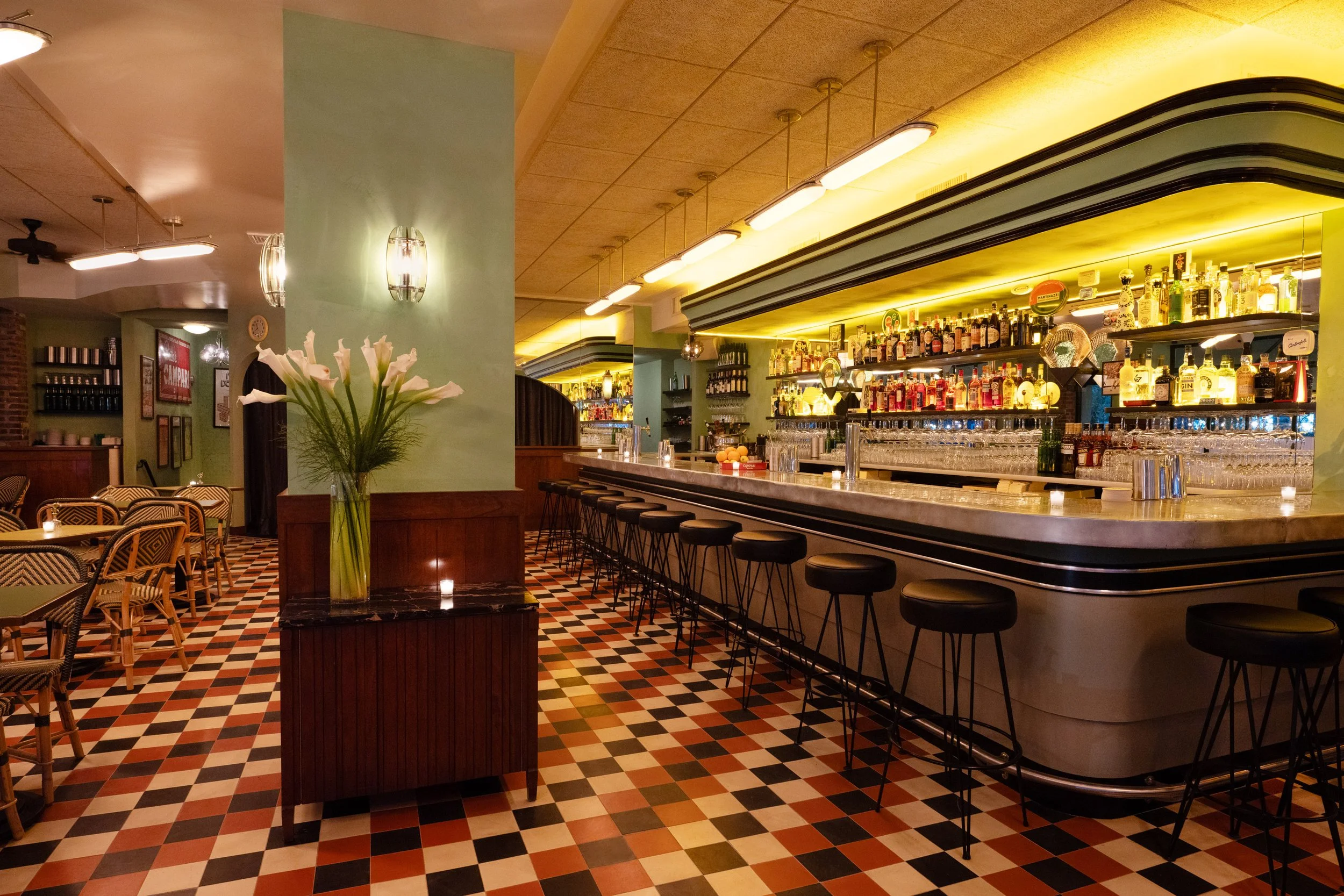 Interior view of a restaurant bar with a colorful checkered floor, green walls, a vase with white flowers, and a long bar counter with stools. Shelves behind the bar display various bottles of alcohol and glassware.