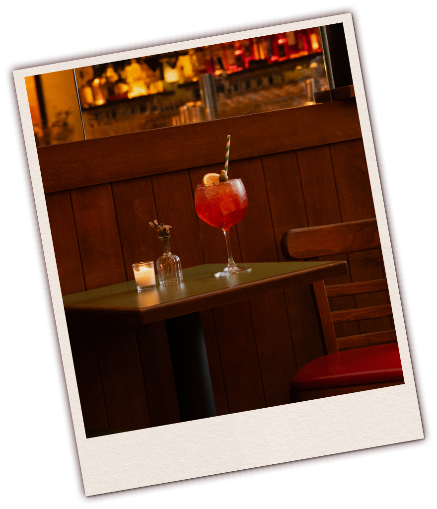 A table with a large red cocktail drink garnished with fruit and a straw, a small candle, and a small vase with flowers, inside a restaurant with warm lighting.