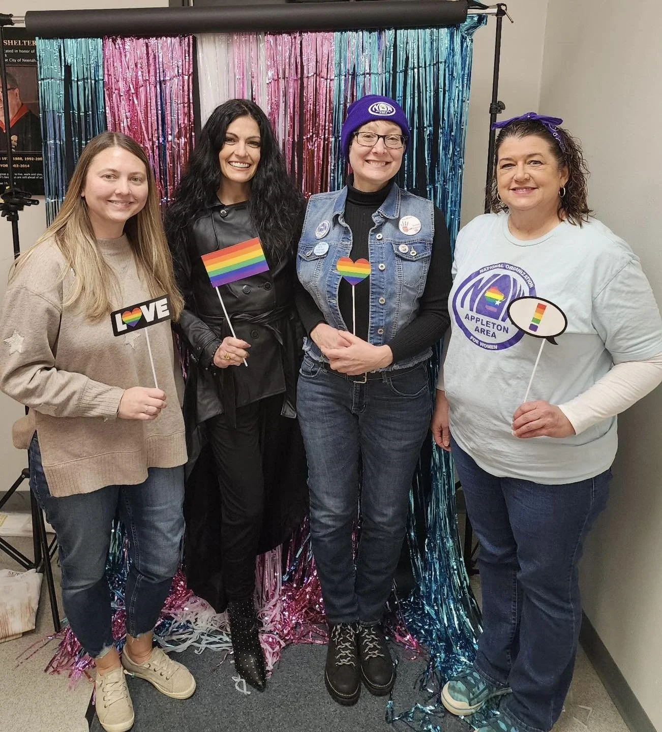 Four women standing together holding rainbow-themed signs in front of colorful tinsel backdrop, smiling.