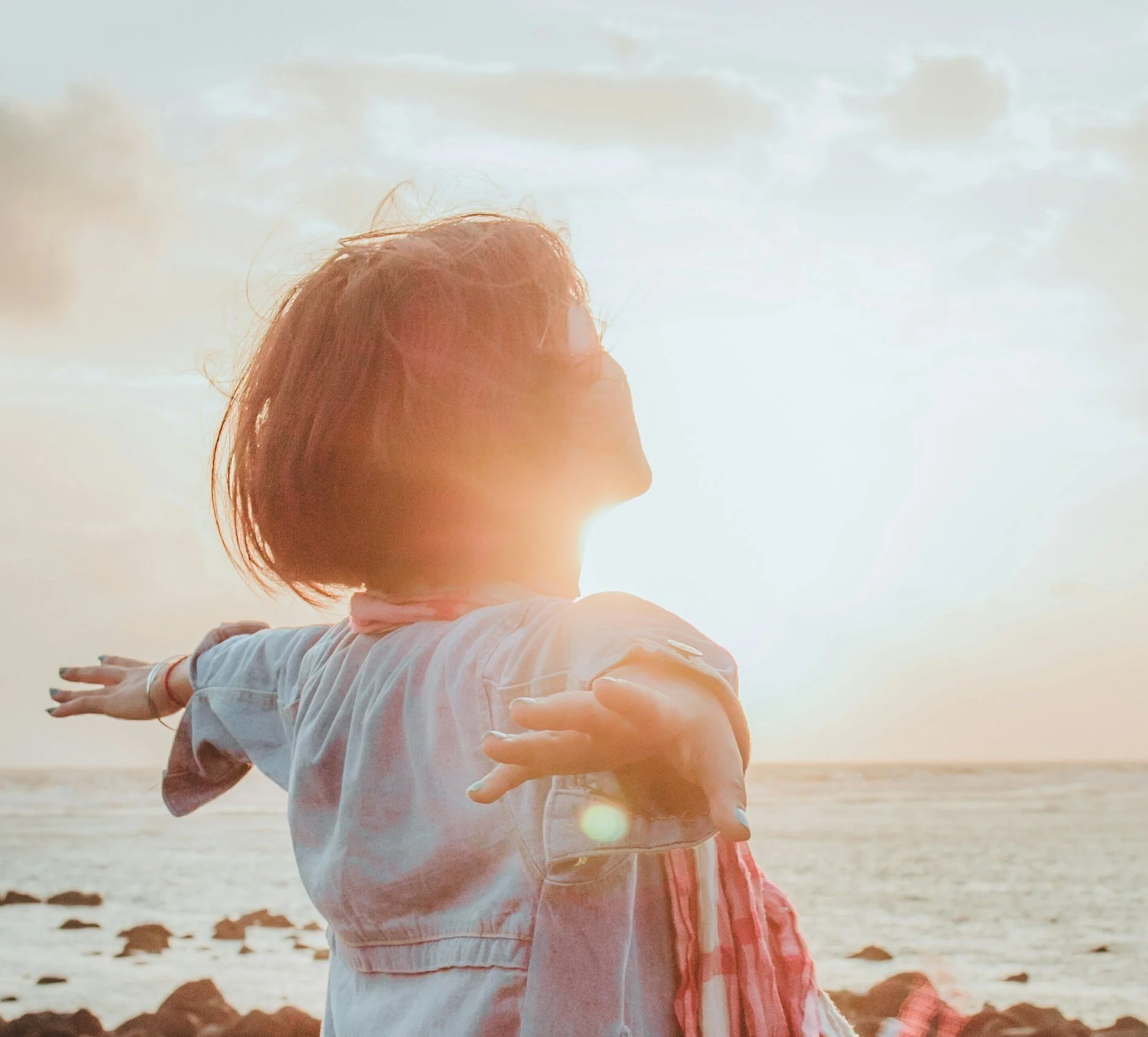 Person with outstretched arms facing the ocean at sunset, bathed in warm sunlight.
