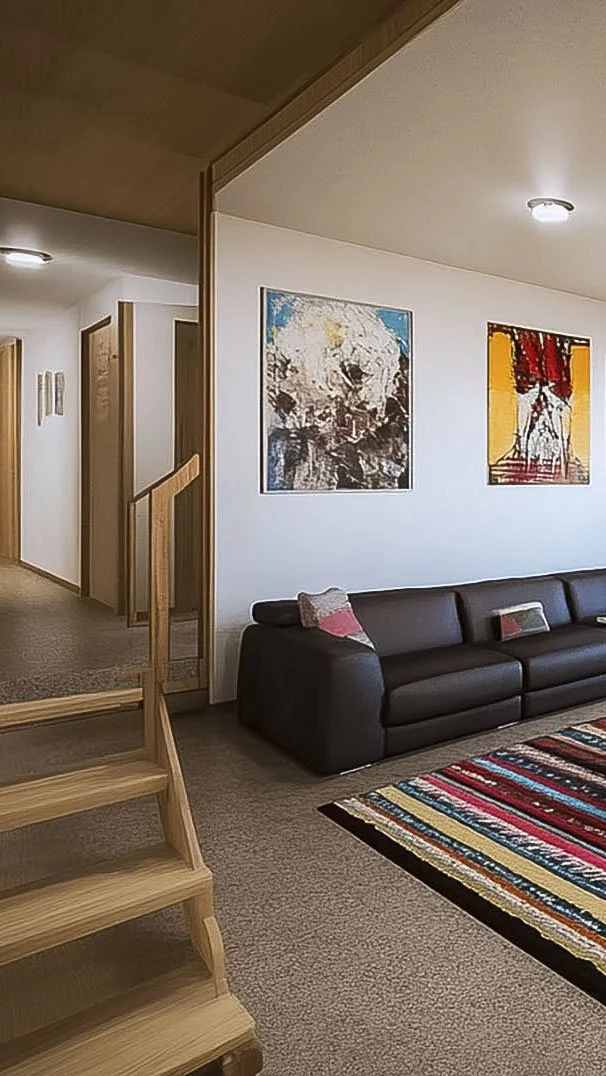 Modern living room interior with a dark gray sofa, colorful striped rug, and abstract art on the wall. Wooden staircase and neutral carpet are visible.