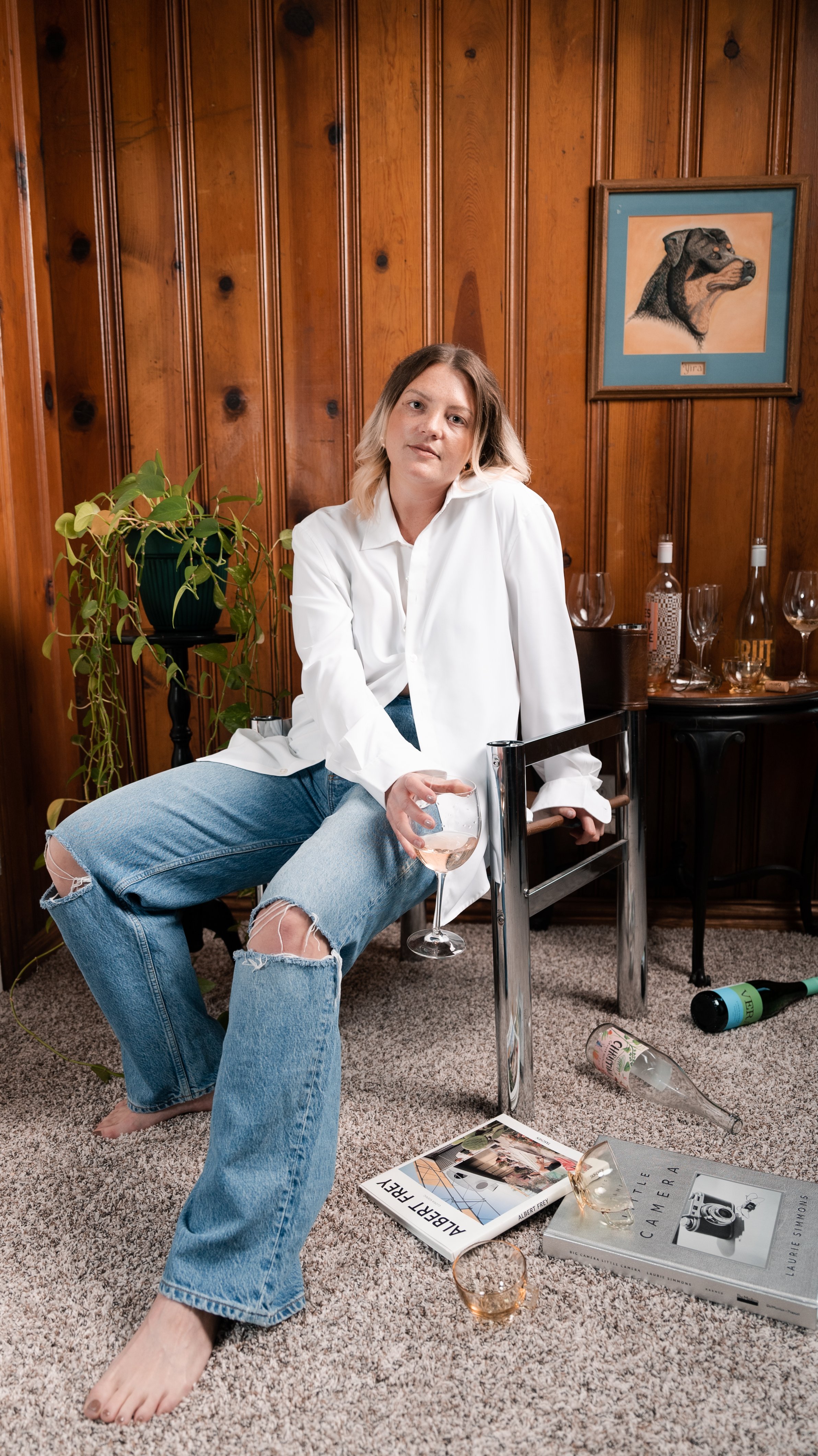 Person sitting on a chair in a wooden-paneled room holding a wine glass, wearing a white shirt and ripped jeans, with books, empty bottles, and glasses on the floor and table.