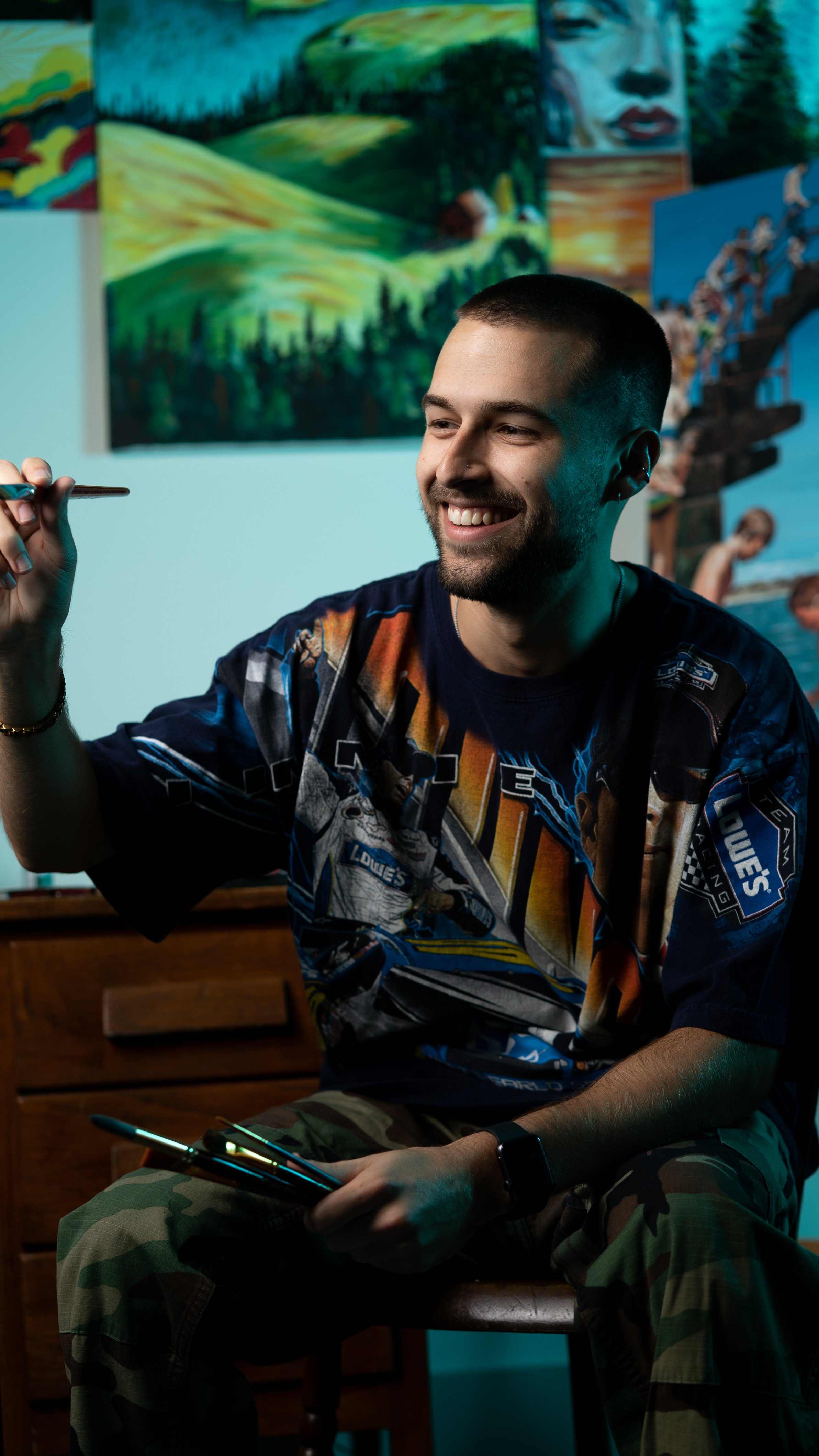 Artist holding a paintbrush, sitting with paintings in the background.