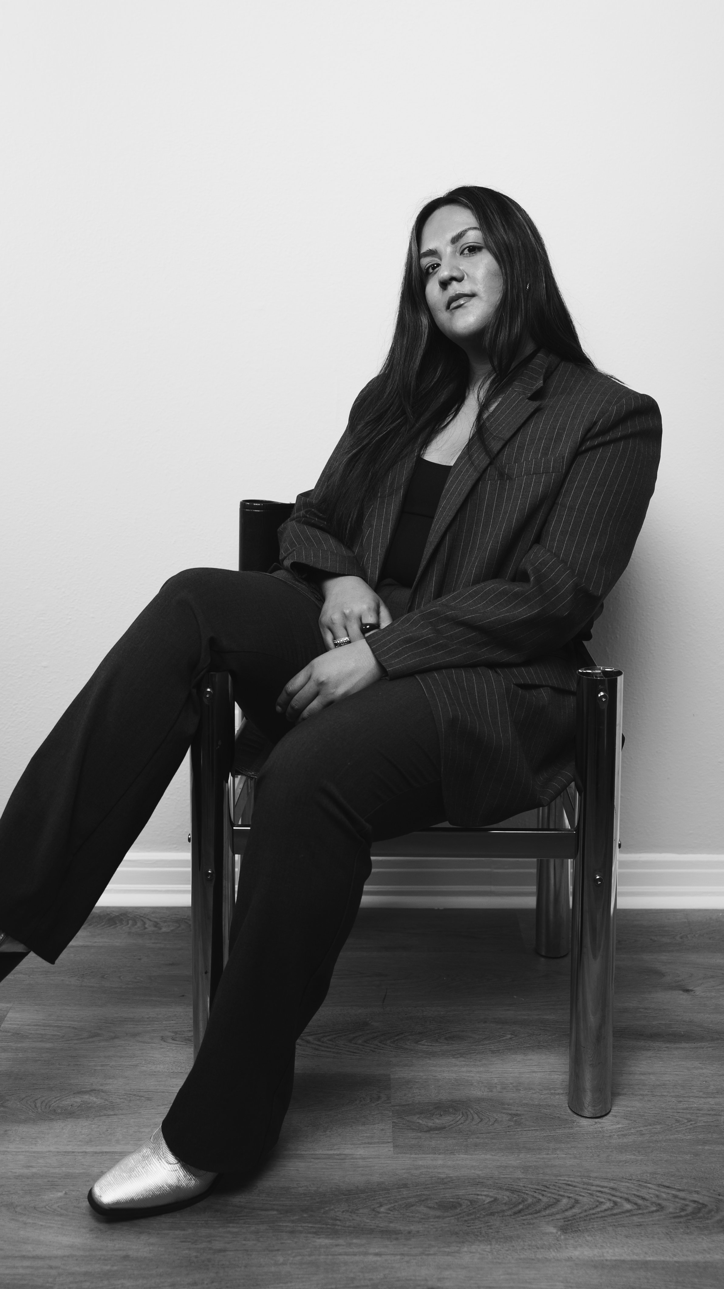 Black and white photo of a person seated in a chair; wearing a pinstripe blazer and dark pants, looking confident, with long hair and silver shoes.