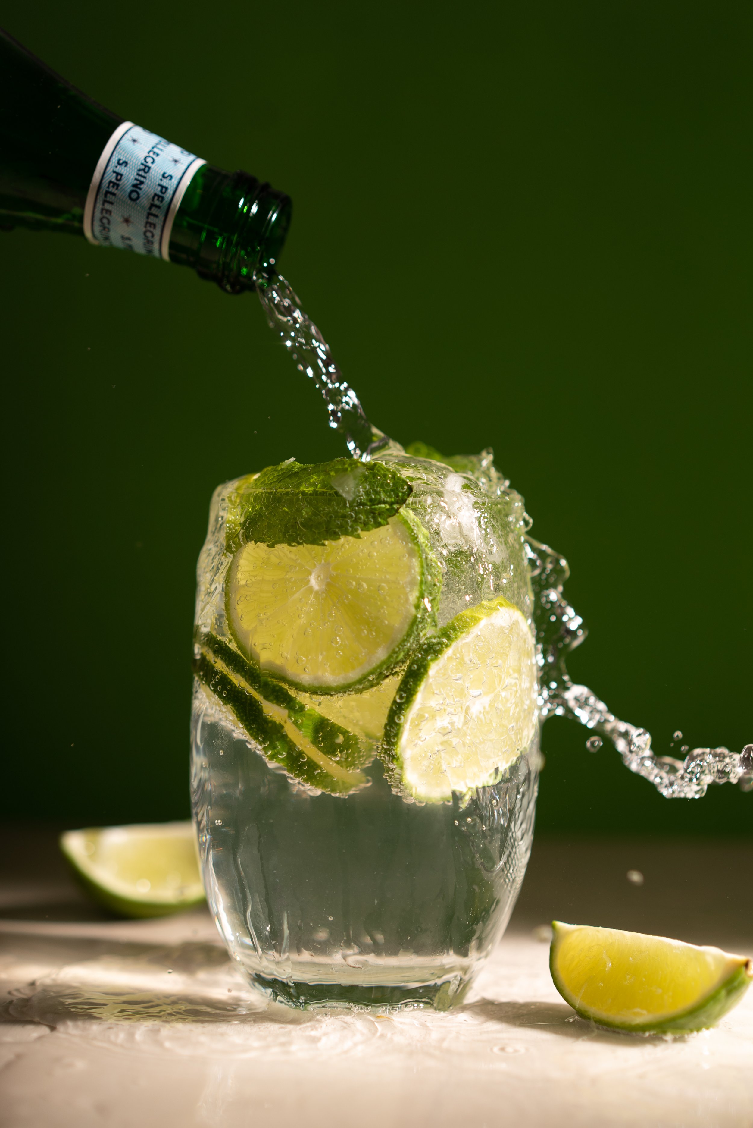 Sparkling water being poured into a glass with lime slices, green background.